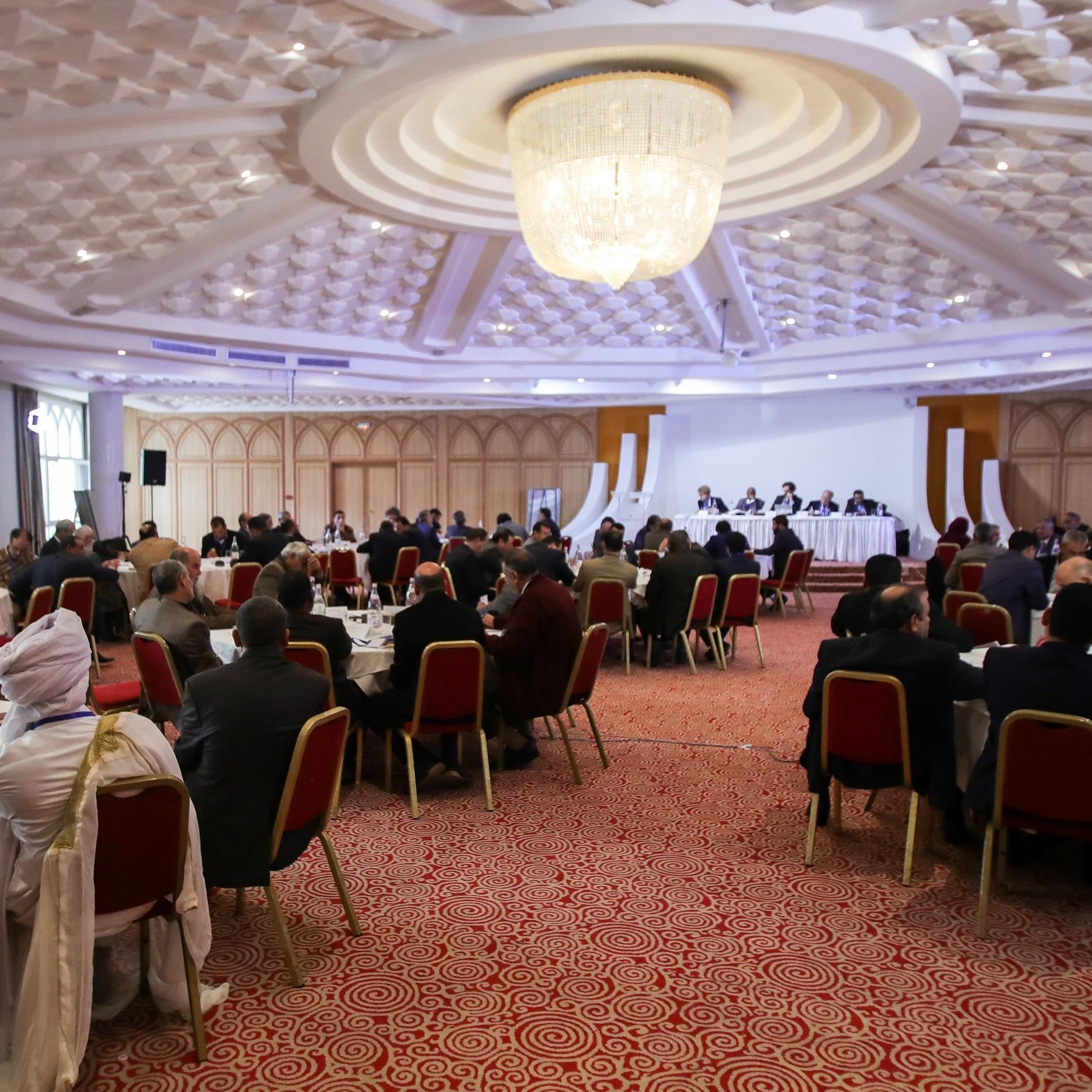 L'image montre une salle de conférence ou de réunion spacieuse, avec de nombreuses personnes assises autour de tables rondes. Les participants semblent engagés dans une discussion ou une présentation, avec un panel sur une estrade à l'avant. La décoration est moderne, avec un plafond orné et un éclairage doux. Les personnes sont habillées de manière formelle, ce qui indique un événement professionnel ou officiel.