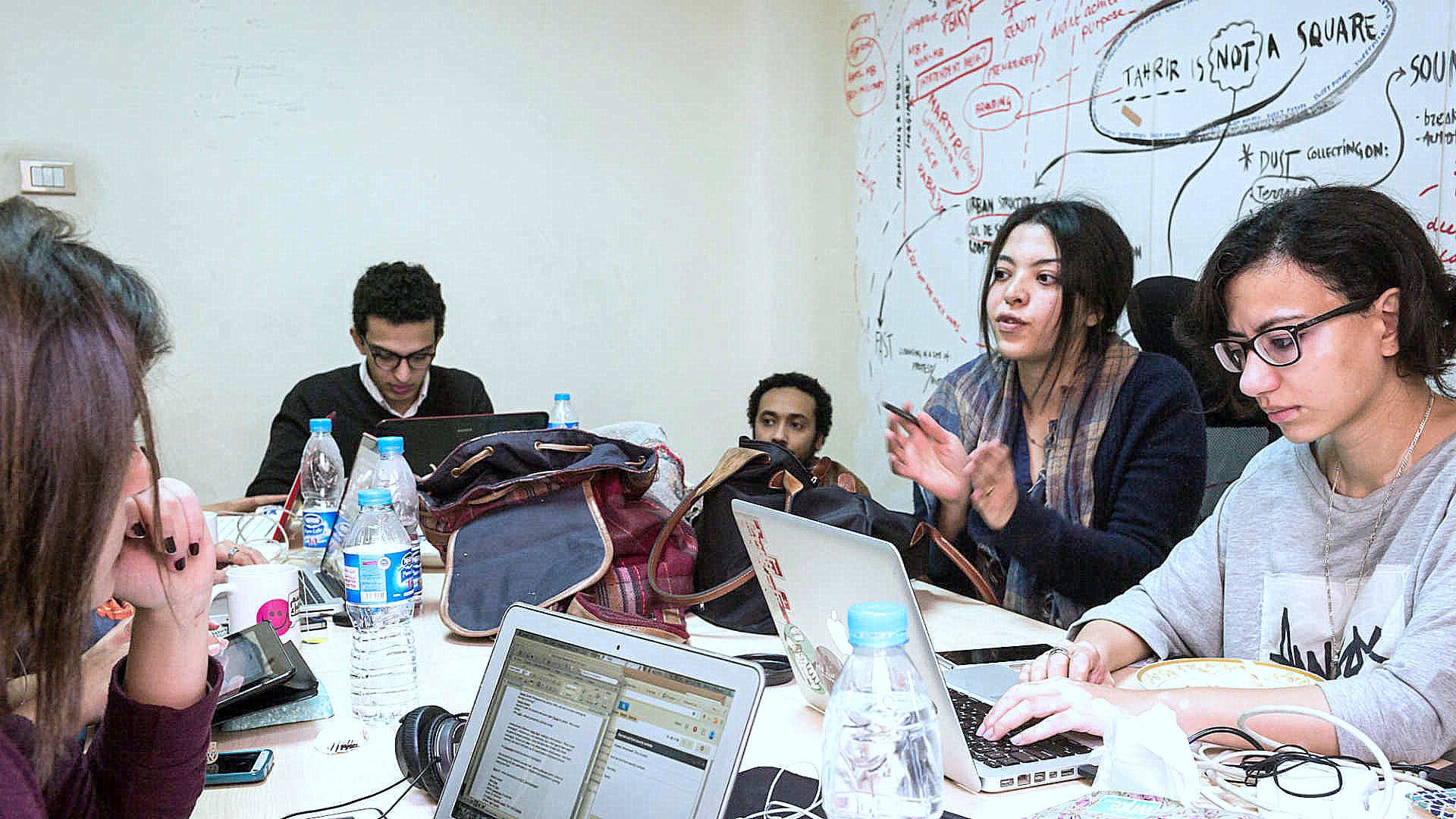 L'image montre un groupe de personnes assises autour d'une table, travaillant sur des ordinateurs portables. Il y a des bouteilles d'eau et des sacs dispersés sur la table. Une femme semble expliquer quelque chose avec enthousiasme, tandis que les autres écoutent attentivement. Le mur derrière eux est couvert de notes et de schémas, suggérant une ambiance créative et collaborative.