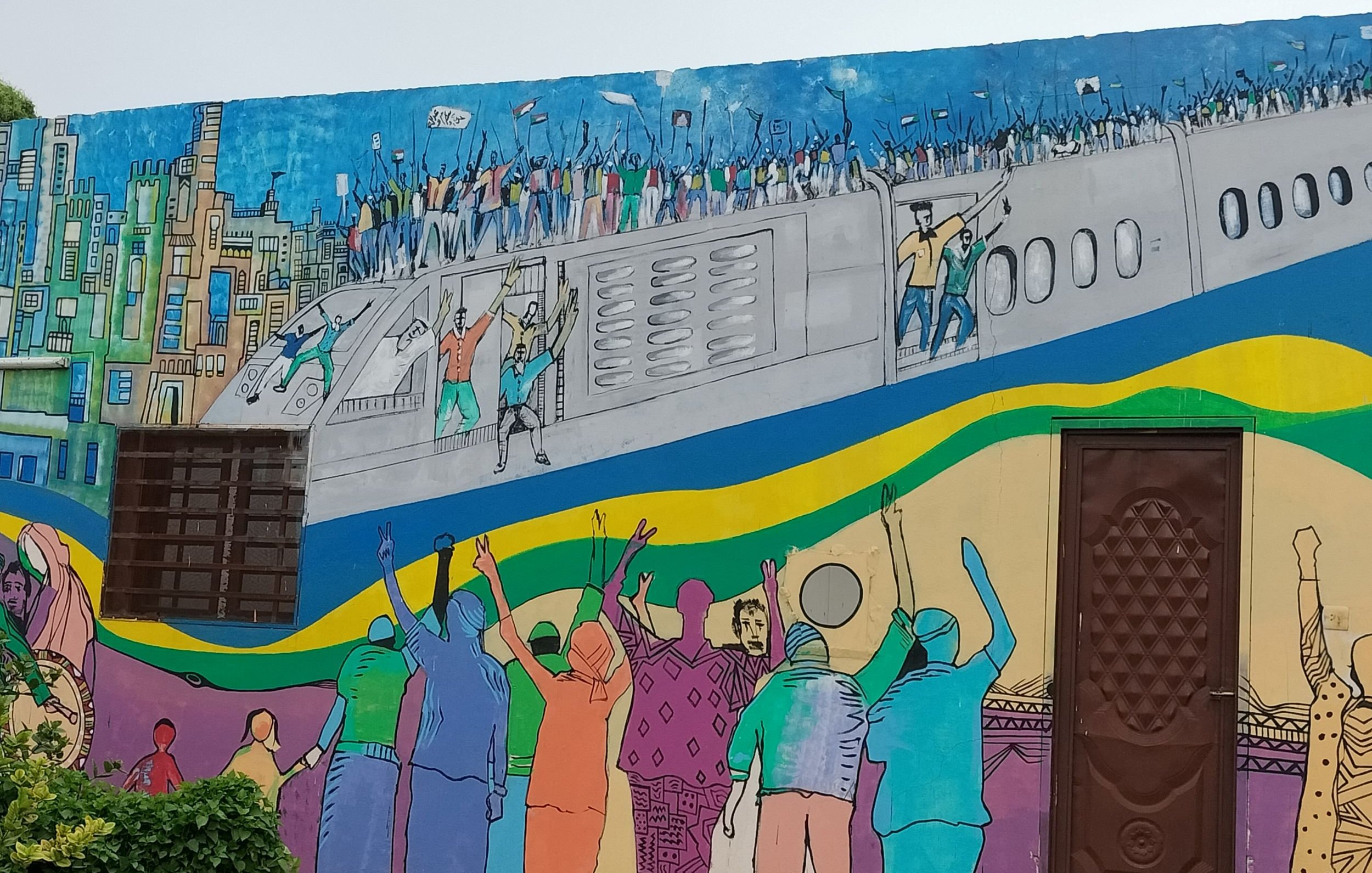 الصورة تظهر جدارية ملونة على حائط، تتضمن مشهدًا حيويًا يبرز مجموعة من الأشخاص الذين يحتفلون أو يتظاهرون. في الجزء العلوي، يظهر مجموعة من الأشخاص على ما يبدو في حالة احتفال، مع أعلام وألوان زاهية. الجزء السفلي يُظهر أشخاصًا يرفعون أيديهم، مما يُعبر عن الحماس أو التأييد. الألوان المستخدمة في الجدارية متنوعة وتجعل الصورة ملفتة للنظر، مما يعطي شعورًا بالحركة والنشاط.