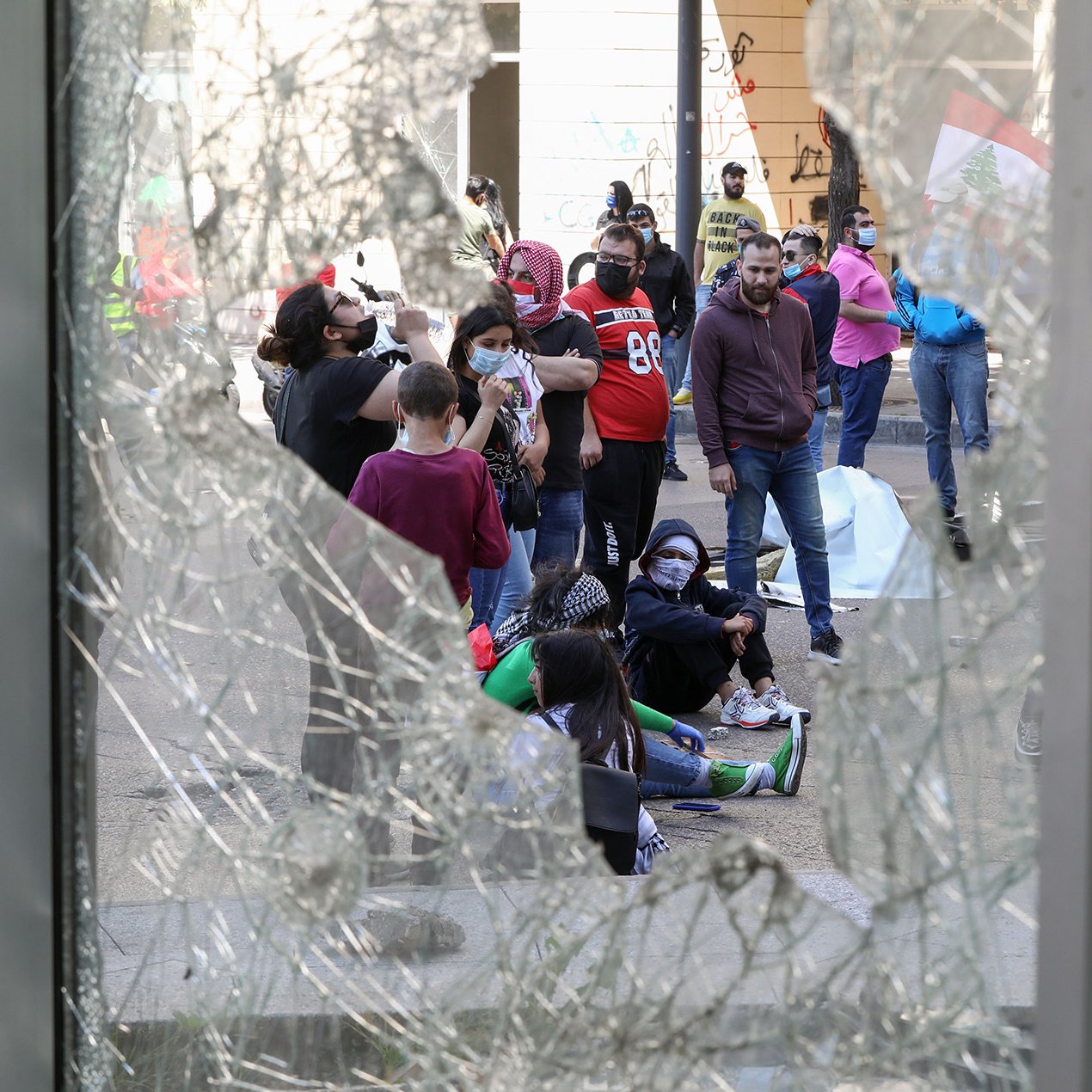 The image shows a group of people gathered on a street, viewed through broken glass. The scene appears to be one of protest or demonstration, as indicated by the presence of individuals wearing masks and the visible graffiti on the walls nearby. Some members of the group are sitting on the ground, suggesting a moment of gathering or reflection amid the chaos. The atmosphere seems tense, reflecting a social or political issue that has drawn this assembly.