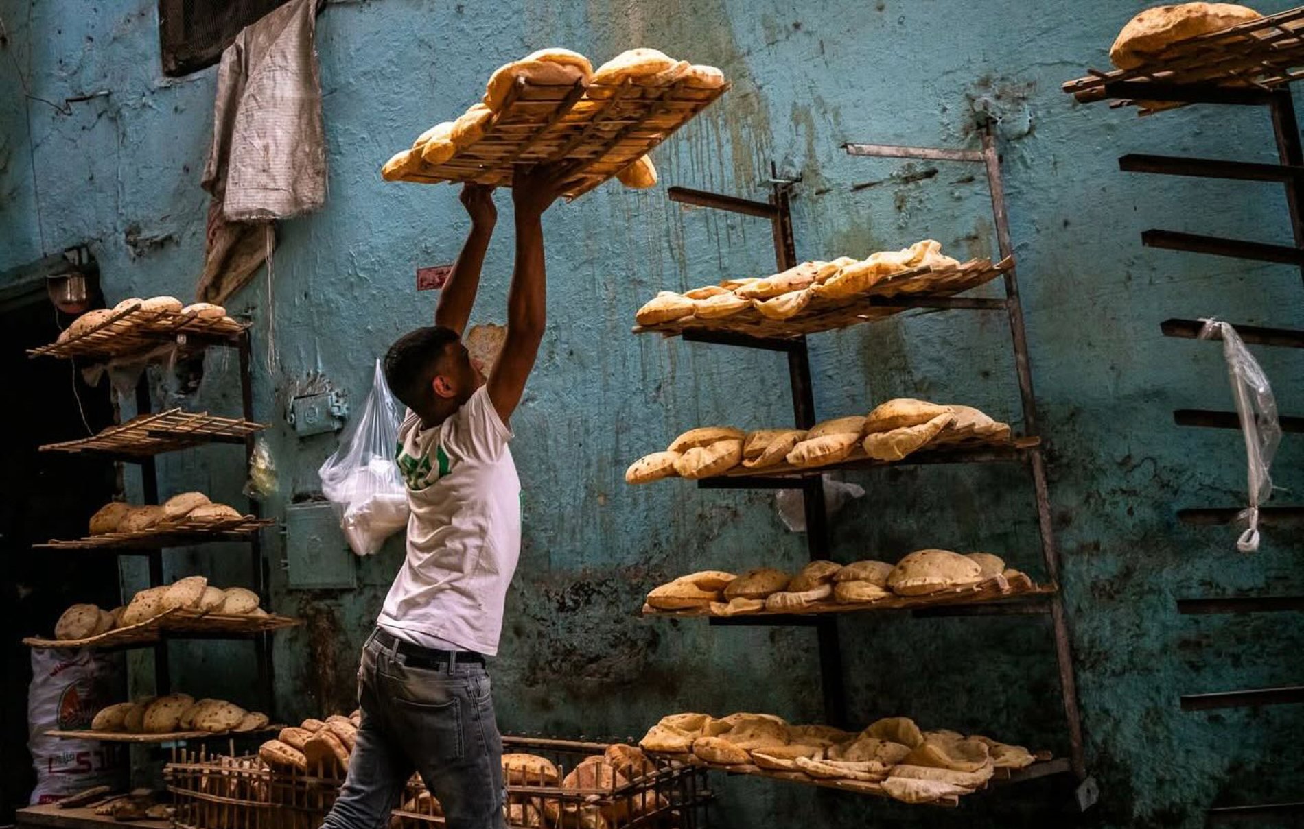 Un boulanger soulève un plateau de pains dans une boulangerie sur un mur bleu.