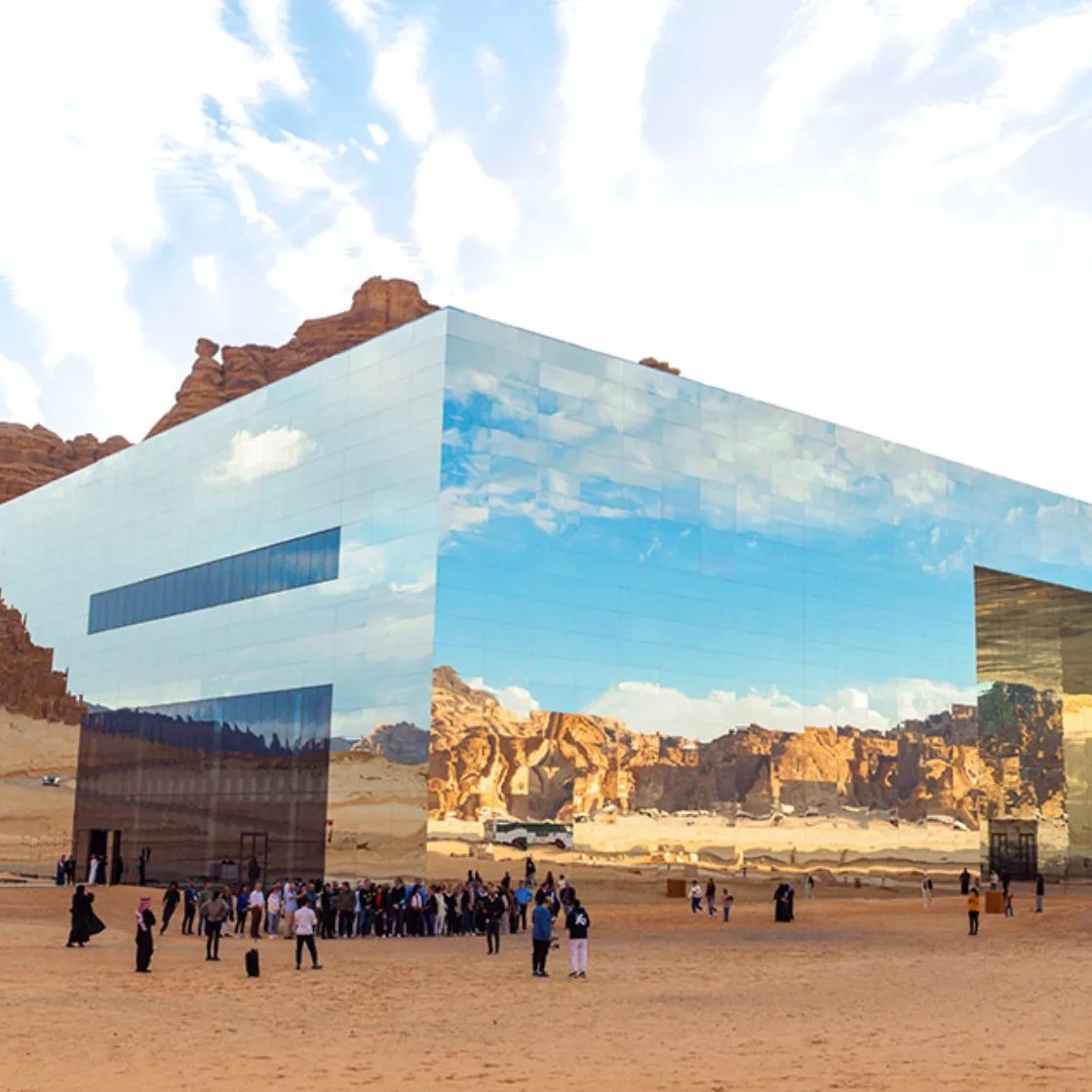 L'image montre un bâtiment moderne et futuriste situé dans un paysage désertique. La structure est entièrement recouverte de miroirs, reflétant le ciel et les montagnes environnantes, ce qui lui permet de se fondre harmoniellement dans le paysage. Au premier plan, un groupe de personnes, vraisemblablement des visiteurs, se regroupe sur le sable. L'atmosphère est paisible, et le ciel est parsemé de nuages, créant un contraste avec les teintes désertiques. L'architecture innovante et les réflexions ajoutent une dimension visuelle fascinante à l'environnement naturel.