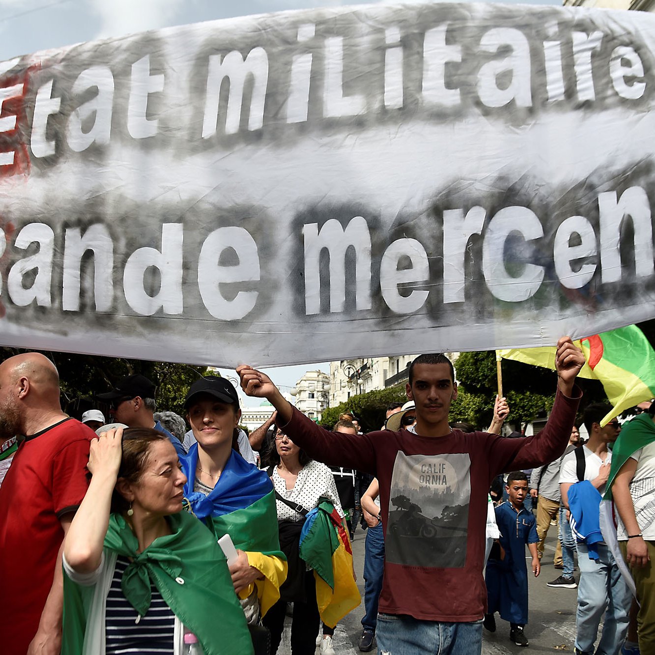 The image shows a large group of people participating in a protest or march. In the foreground, a man is holding a prominent banner that reads "Ni Etat Militaire Ni Bande Mercenaire," which translates to "Neither Military State Nor Mercenary Band." The crowd appears diverse, with individuals carrying flags and wearing various clothing styles, indicative of a collective demonstration. The background features urban scenery, suggesting the protest is taking place in a city.