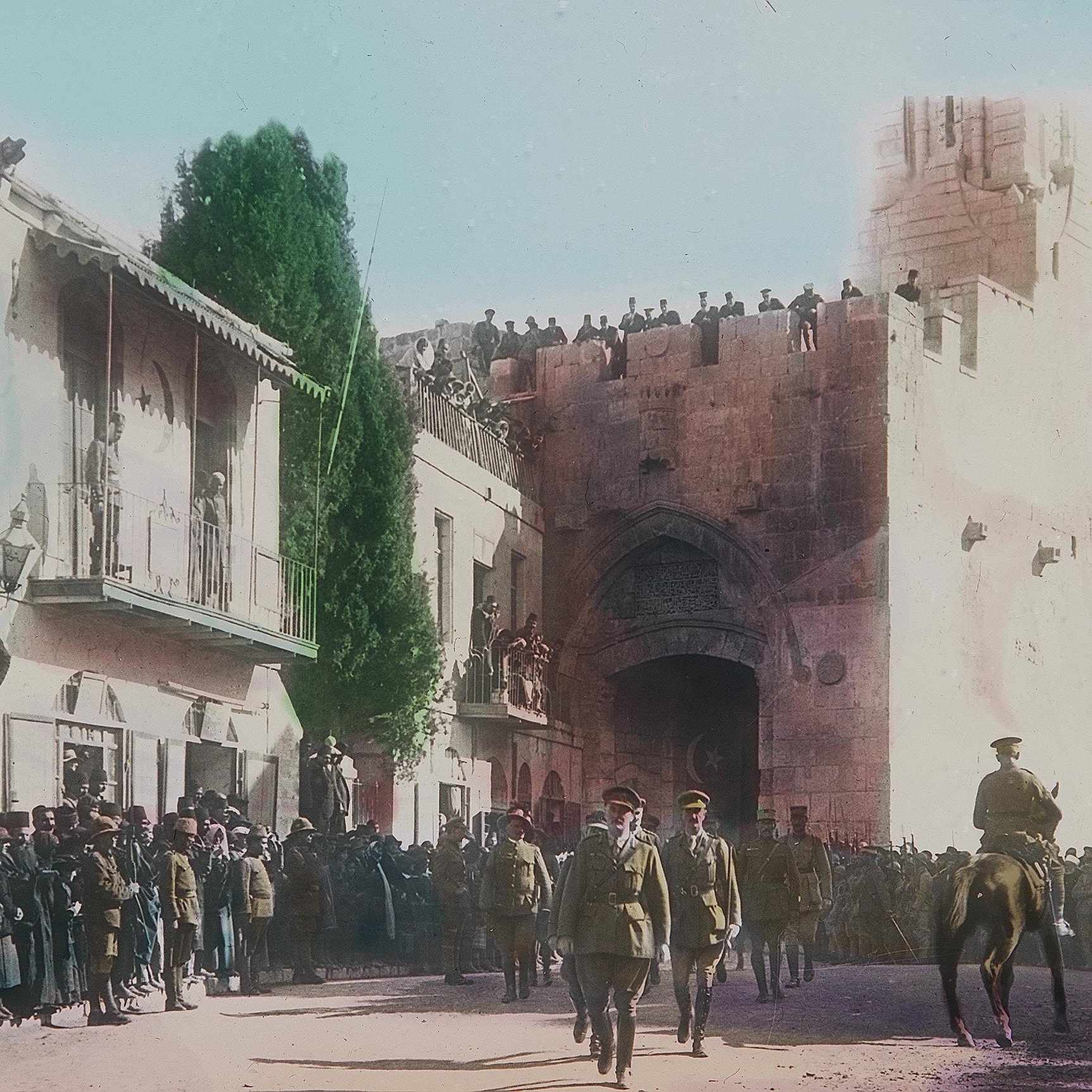 Cette image, en couleur, montre une scène historique se déroulant probablement lors d'un événement important à Jérusalem. Au premier plan, des soldats en uniforme, dont certains à cheval, avancent avec une certaine autorité. En arrière-plan, il y a une porte majestueuse sur un mur ancien, entourée de personnes qui regardent la procession depuis des balcons et des hauteurs. La foule est dense, avec des hommes, des femmes et des enfants, tous attentifs à ce qui se passe. Les bâtiments environnants sont typiques de l'architecture de la région, et l'atmosphère semble être à la fois festive et solennelle.