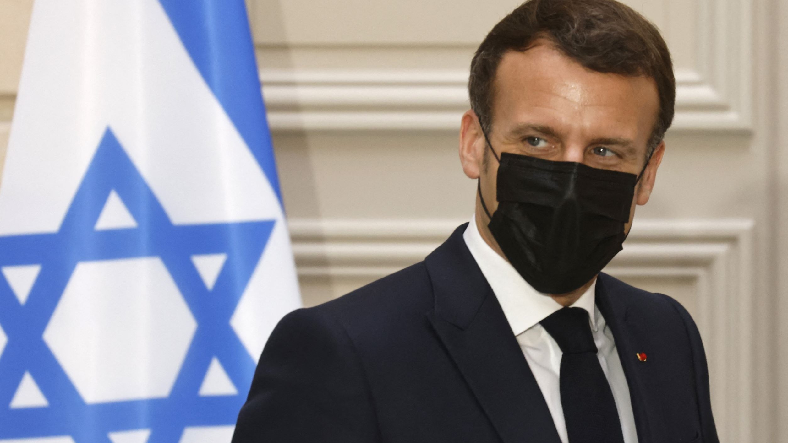 L'image montre une personne en costume, portant un masque noir, se tenant près d'un drapeau qui comporte une étoile de David. L'arrière-plan semble être un intérieur formel, peut-être un bureau ou une salle de réunion. La personne a une expression attentive et semble engagée dans une conversation.