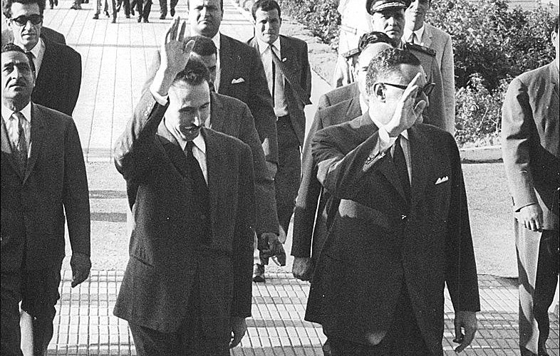 The image depicts a group of men walking together outdoors in a formal setting. Two men in the foreground are waving, suggesting they are acknowledging an audience or photographers. They are dressed in suits, indicating a formal occasion. Behind them, several other men in suits and uniforms follow, along with some individuals in lighter clothing, possibly indicating a recent gathering or event. The background features greenery and is well-organized, contributing to a dignified atmosphere.