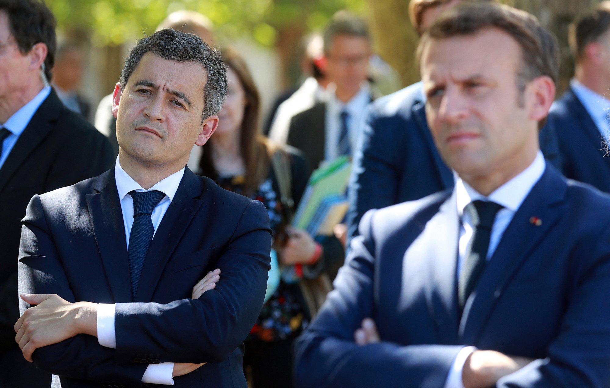 L'image montre deux hommes en costume, l'un avec les bras croisés et l'air pensif, et l'autre légèrement en avant, observant. Ils semblent participent à un événement public, entourés d'autres personnes en arrière-plan, dans un cadre extérieur. Les expressions faciales suggèrent qu'ils écoutent attentivement ou réfléchissent à ce qui se passe.