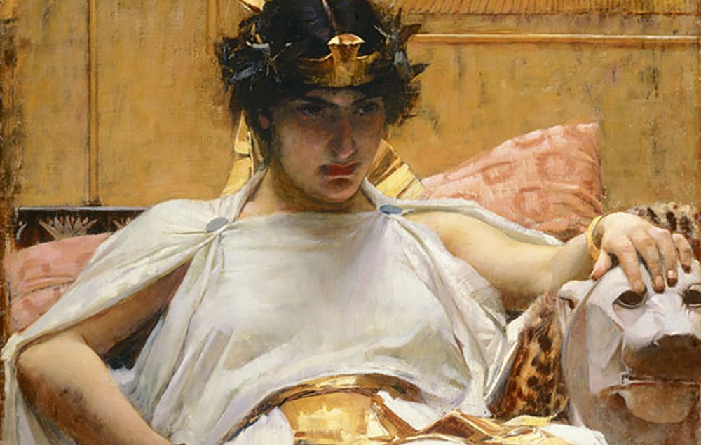 L'image représente une femme assise sur un siège orné, drapée dans une tunique blanche et une écharpe dorée. Elle porte une couronne et a les cheveux sombres, avec un regard pensif et sérieux. À côté d'elle, on aperçoit un lion stylisé sur le siège. L'arrière-plan est décoré de motifs égyptiens, avec des couleurs chaudes et une ambiance luxueuse. La composition évoque une atmosphère royale et mystique.