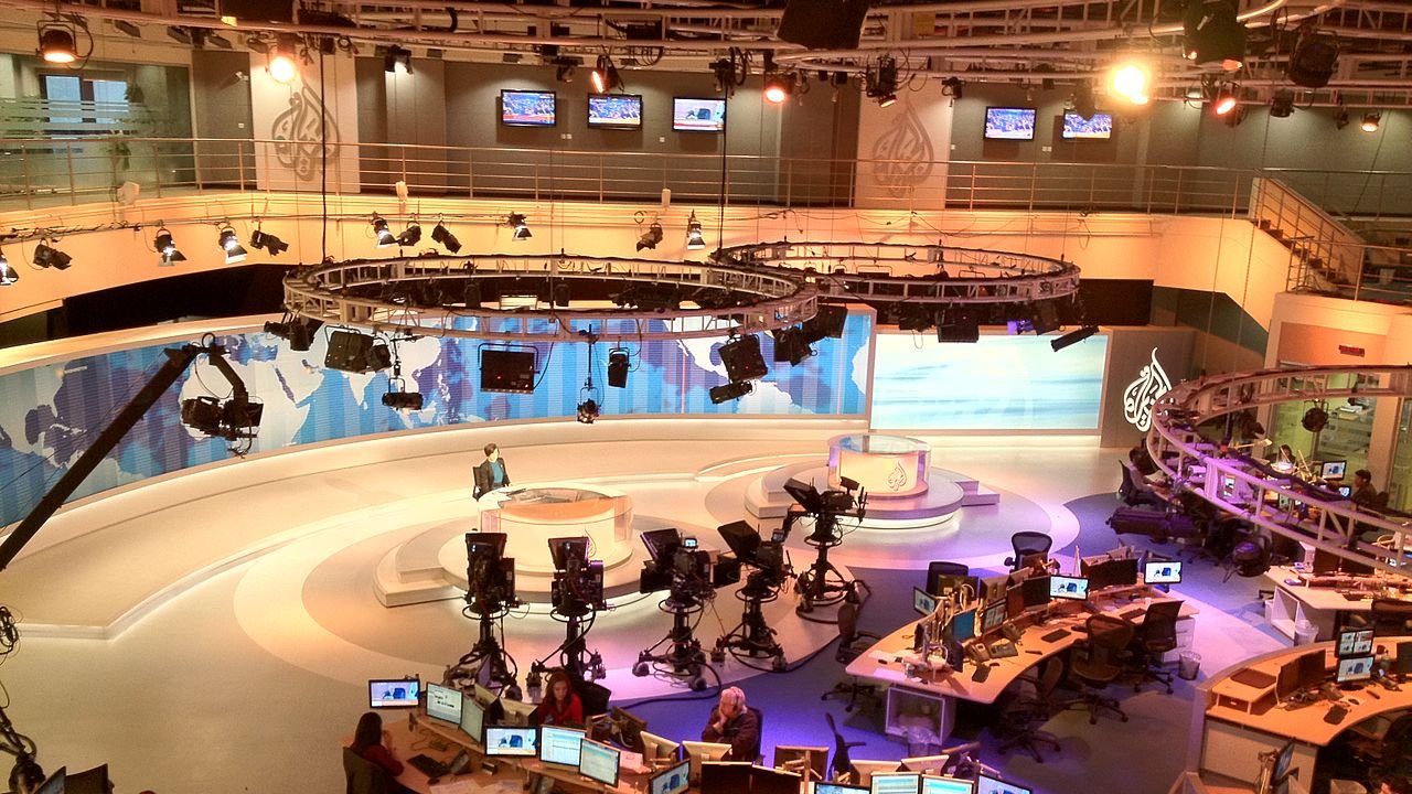 L'image montre un studio de télévision moderne, probablement utilisé pour l'information. On y voit plusieurs caméras placées sur des trépieds, ainsi que des moniteurs et des bureaux avec des ordinateurs. Le décor est élégant, avec des panneaux de fond aux couleurs douces et des éclairages suspendus. Au centre, il y a un espace pour le présentateur, entouré d'une atmosphère professionnelle et technologique. L'ensemble donne une impression de dynamisme et de préparation pour diffuser des nouvelles en direct.