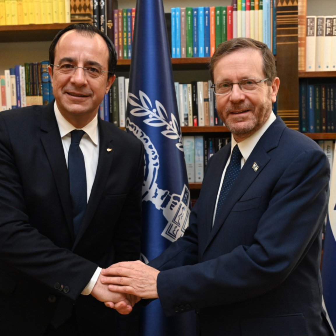 Deux hommes en costume se serrent la main devant des drapeaux et des étagères de livres.