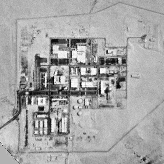 L'image montre une vue aérienne en noir et blanc d'une installation industrielle ou militaire située dans une zone désertique. On peut y voir plusieurs bâtiments et structures disposés de manière ordonnée, entourés d'une clôture ou d'une barrière. Le paysage environnant semble aride, avec peu de végétation visible. Cette configuration suggère un site ayant des activités techniques ou de recherche avancées.