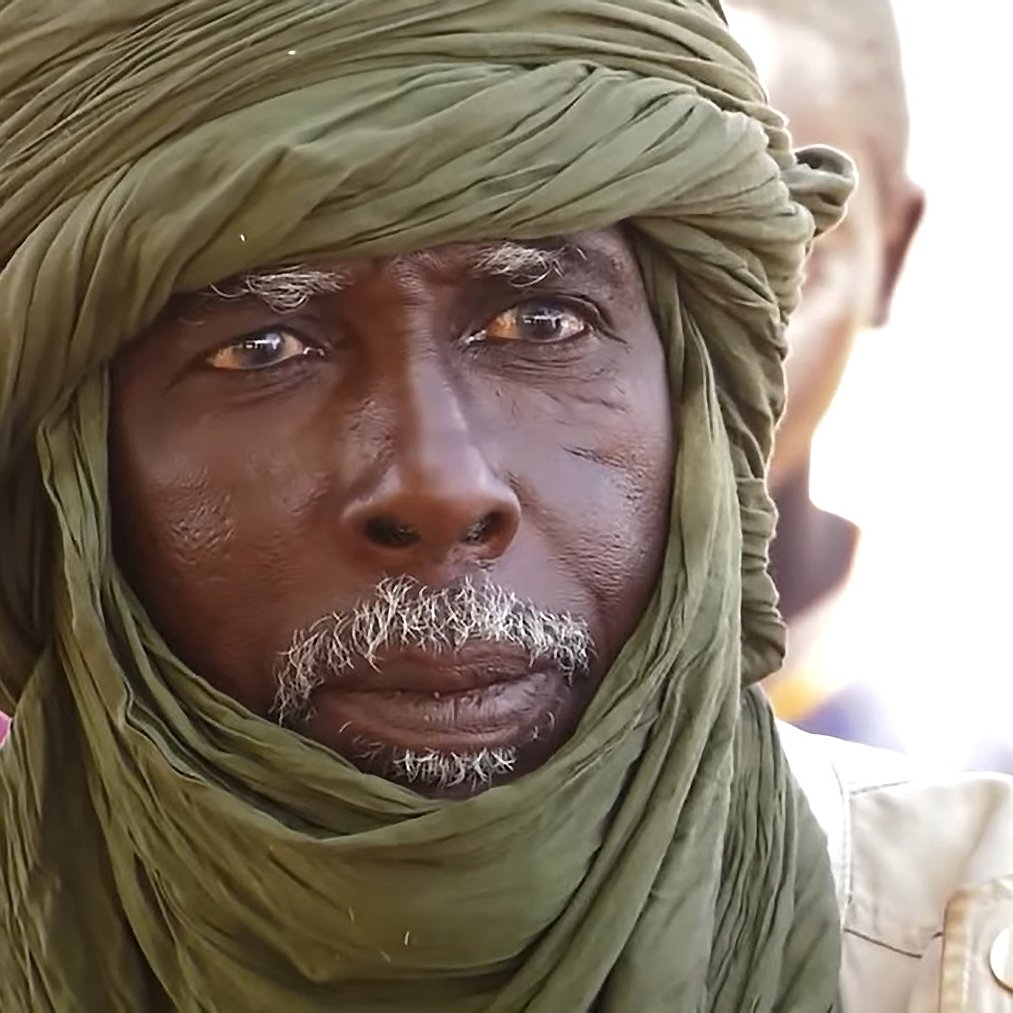 The image features an elderly man with a distinguished appearance. He is wearing a green turban wrapped around his head, which adds to his dignified look. His face shows signs of age, with deep wrinkles and a white beard, suggesting wisdom and experience. The background appears to be a social setting with other individuals present, though they are not clearly defined in the image. The focus is on the man, capturing a solemn expression that hints at deep thought or contemplation.