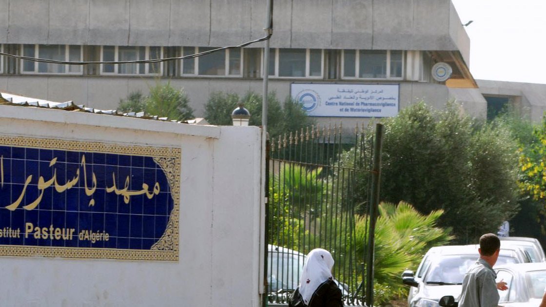 The image depicts a building with signage indicating it is the "Institut Pasteur" in Algeria. In the foreground, there is a decorative wall with Arabic text, likely the name of the institution, along with some greenery. You can also see several people walking along a path near the entrance. The architectural style of the building appears modern and utilitarian.