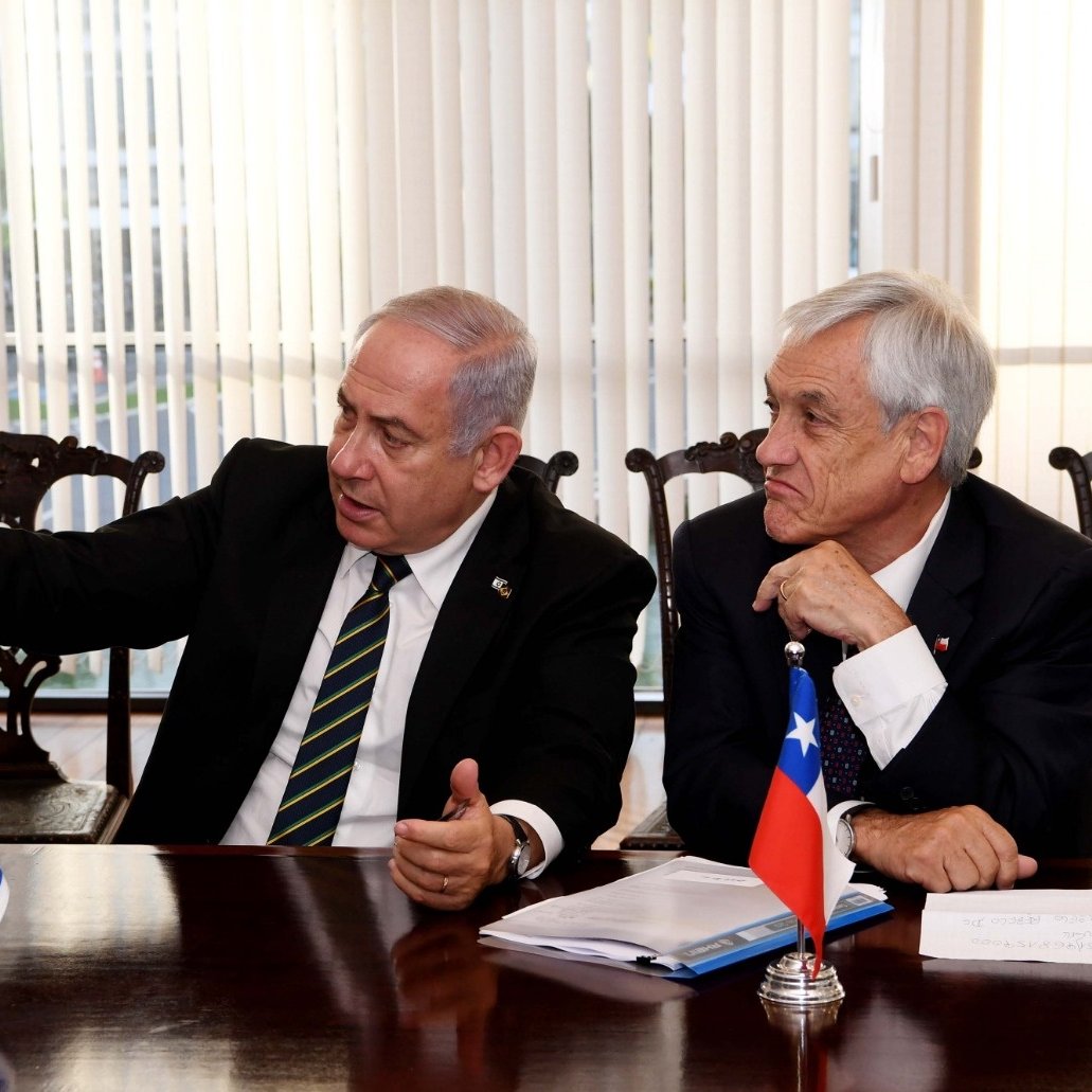 L'image montre deux hommes assis à une table. L'un d'eux, à gauche, gesticule en parlant, tandis que l'autre écoute attentivement. Sur la table se trouvent des documents, et des drapeaux d'Israël et du Chili sont visibles à côté d'eux. L'atmosphère semble sérieuse, suggérant une discussion diplomatique ou politique. Les rideaux derrière donnent une ambiance lumineuse à la scène.