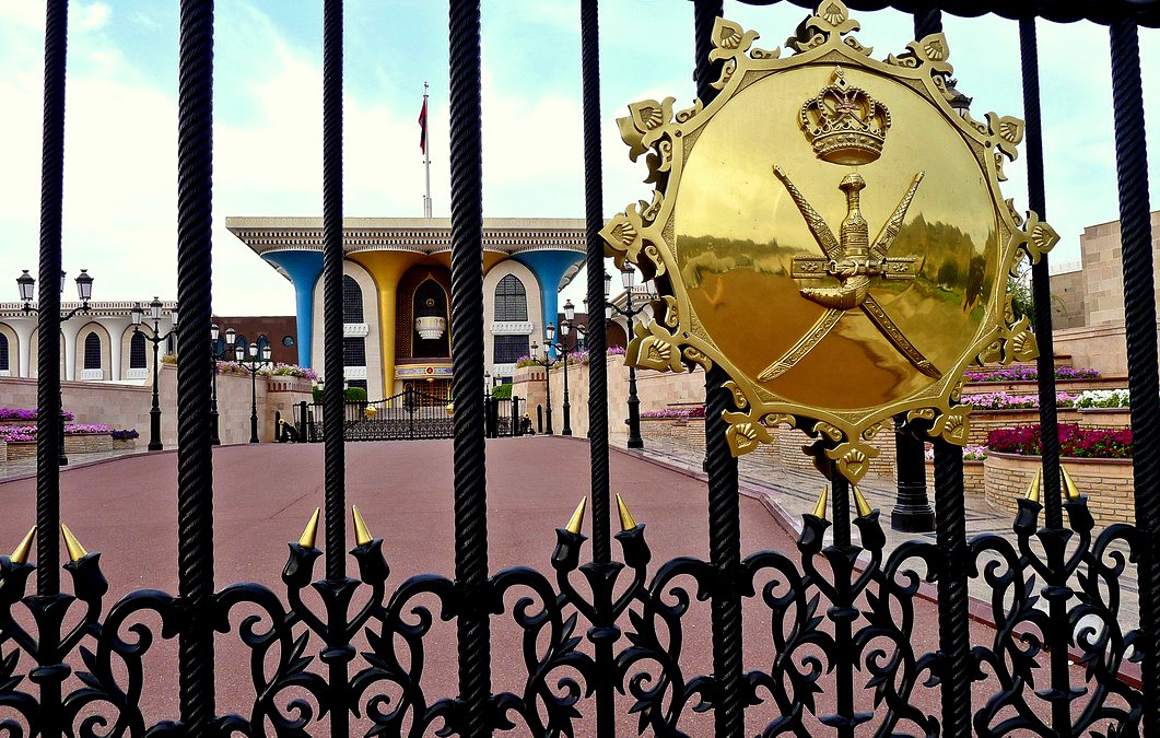 L'image montre un portail d'entrée orné de motifs métalliques élaborés. Au centre, on aperçoit un blason doré avec une couronne et des épées croisées, symbolisant probablement un gouvernement ou une monarchie. À l'arrière-plan, on distingue un bâtiment aux couleurs claires et un drapeau flottant, entouré de pelouses et de fleurs. L'atmosphère dégage une sensation de grandeur et d'autorité.