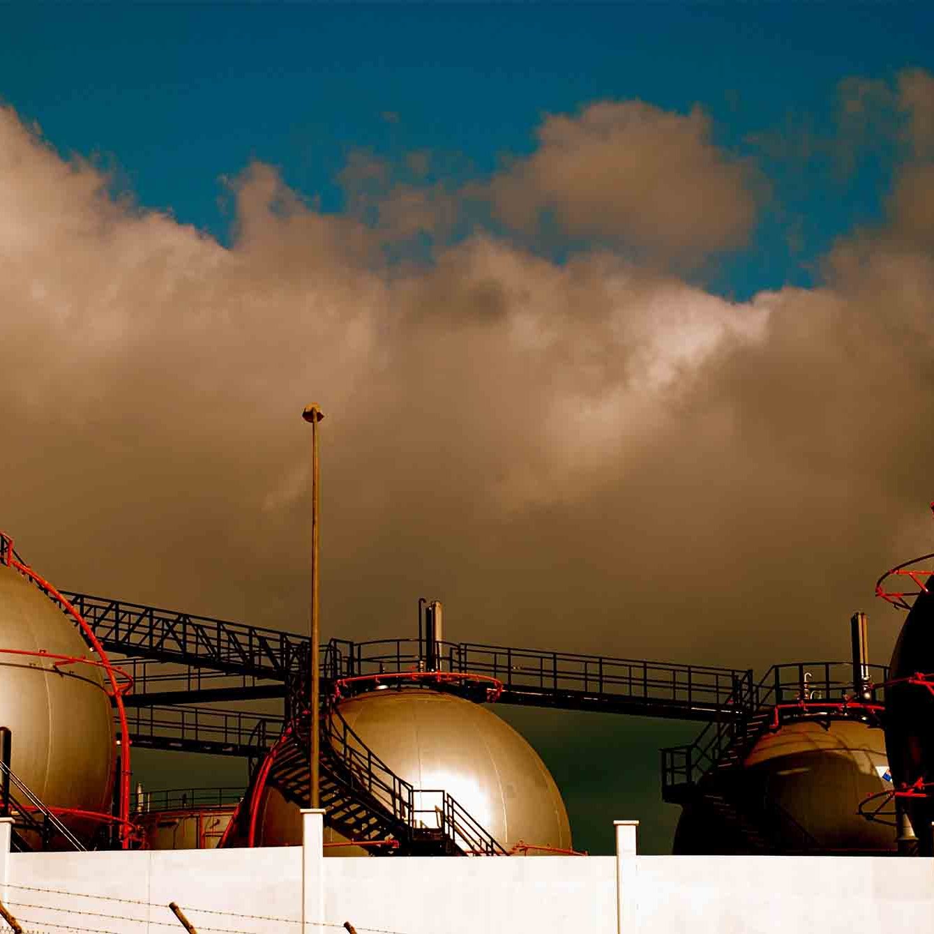 L'image présente des réservoirs à gaz de forme sphérique, surmontés de structures métalliques. Ils sont disposés sur une plateforme, tandis qu'un ciel nuageux et dramatique se profile en arrière-plan. Les couleurs à la fois sombres et lumineuses créent une atmosphère industrielle marquée par le contraste entre les réservoirs et le ciel.