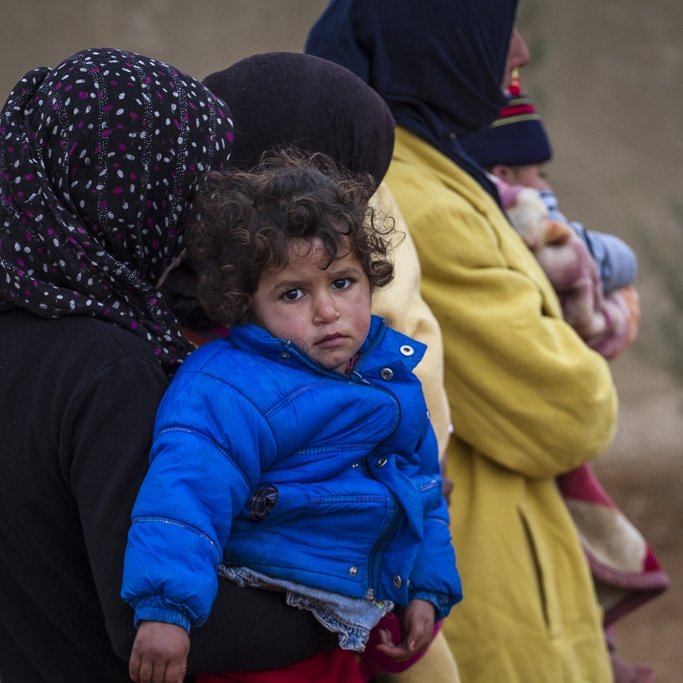 L'image montre plusieurs personnes, dont une femme tenant un enfant dans ses bras. L'enfant porte une veste bleue, et son expression semble triste ou préoccupée. Les femmes qui les entourent sont habillées de manière chaleureuse et portent des foulards. L'arrière-plan est flou, avec des éléments naturels, ce qui suggère un environnement difficile. L'ensemble de la scène évoque une atmosphère de vulnérabilité et de défi.