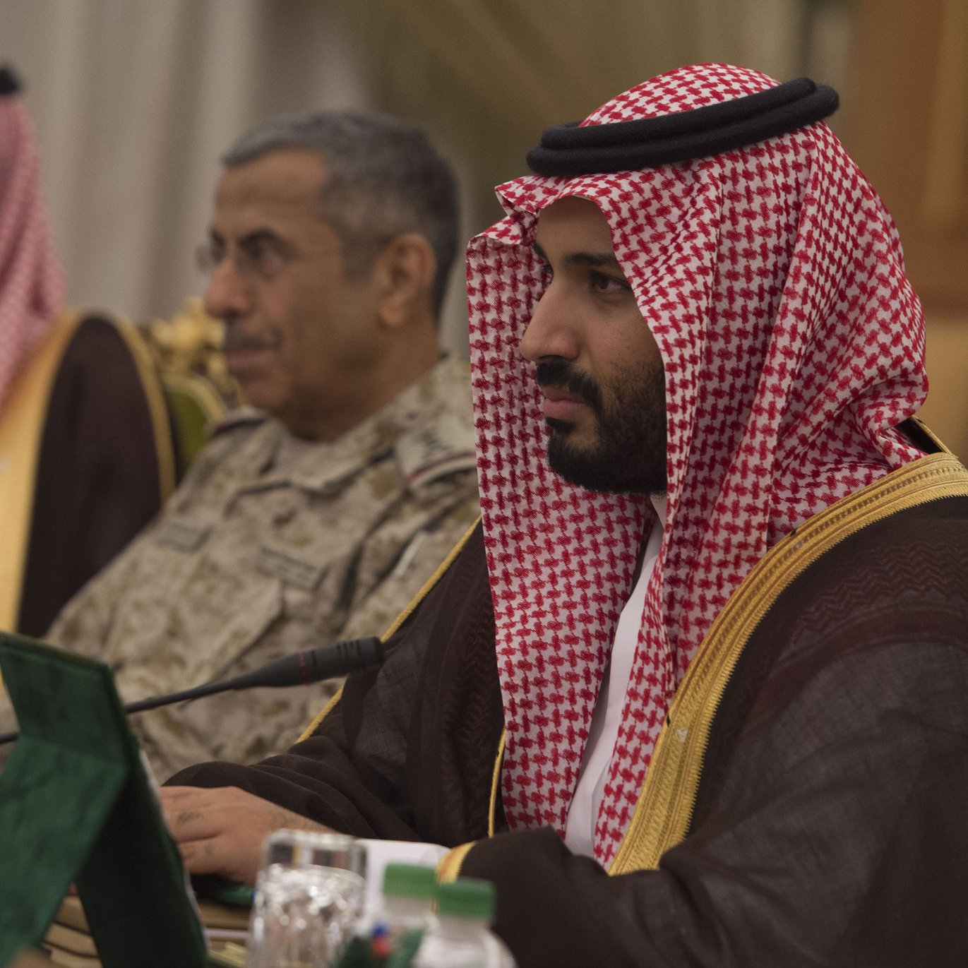 L'image montre un groupe de personnes lors d'une réunion. Au premier plan, un homme porte une tenue traditionnelle saoudienne, incluant un headdress appelé ghutrah ou agal. Il semble pensif et attentive. À l'arrière-plan, un autre homme en uniforme militaire est également présent, écoutant la discussion. Des drapeaux et des documents sont visibles sur la table, suggérant une réunion officielle ou diplomatique. L'environnement a un air formel et sérieux, propice aux échanges importants.