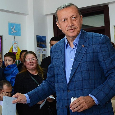 L'image montre un homme d'âge moyen portant un costume bleu. Il est debout, tenant un bulletin dans sa main, et semble être dans une salle de vote ou un endroit similaire. Il y a plusieurs personnes en arrière-plan, dont une femme avec des lunettes, un enfant et d'autres adultes, qui regardent l'homme avec intérêt. L'environnement est chaleureux et coloré, avec des affiches accrochées aux murs.