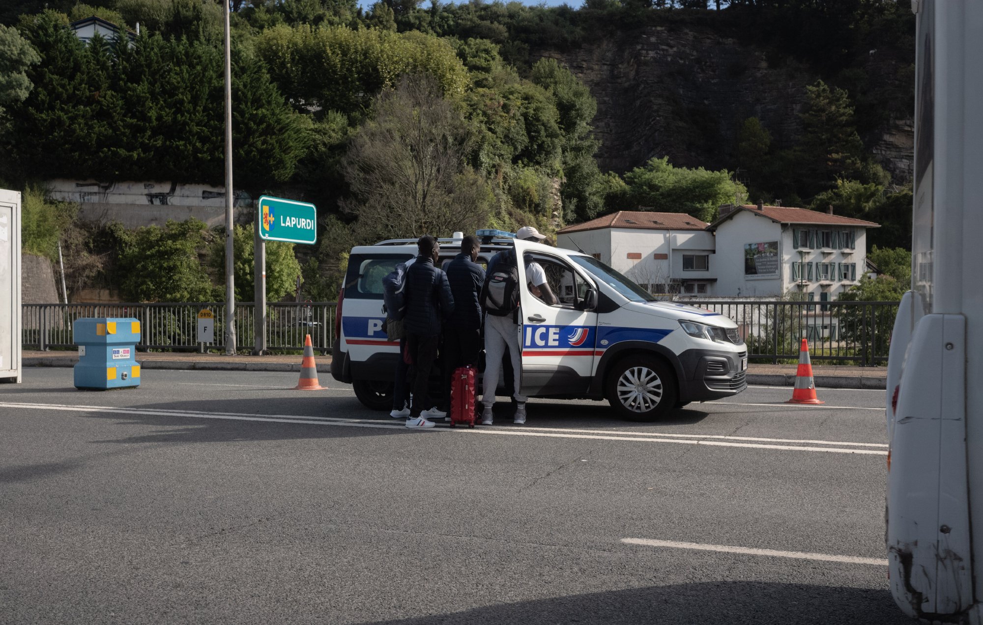 The image shows a scene on a road where a police vehicle is stopped. A few individuals are standing near the back of the police car, which is marked with the word "Police." The setting appears to be a highway or a major road, with some road cones placed nearby. In the background, there are trees and some buildings visible, suggesting an urban or suburban area. The atmosphere seems to indicate a police check or an interaction taking place.