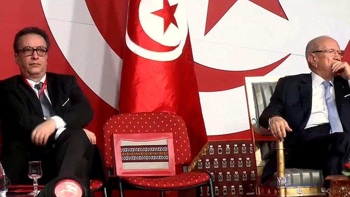 L'image montre deux hommes assis sur des chaises lors d'un événement. Ils semblent être en train d'écouter ou d'observer quelque chose, peut-être un discours ou une présentation. En arrière-plan, il y a un drapeau tunisien, ce qui indique que l'événement se déroule en Tunisie. L'un des hommes porte des lunettes et a une expression sérieuse, tandis que l'autre exerce une posture détendue. La scène semble officielle, peut-être liée à la politique ou à un rassemblement important.