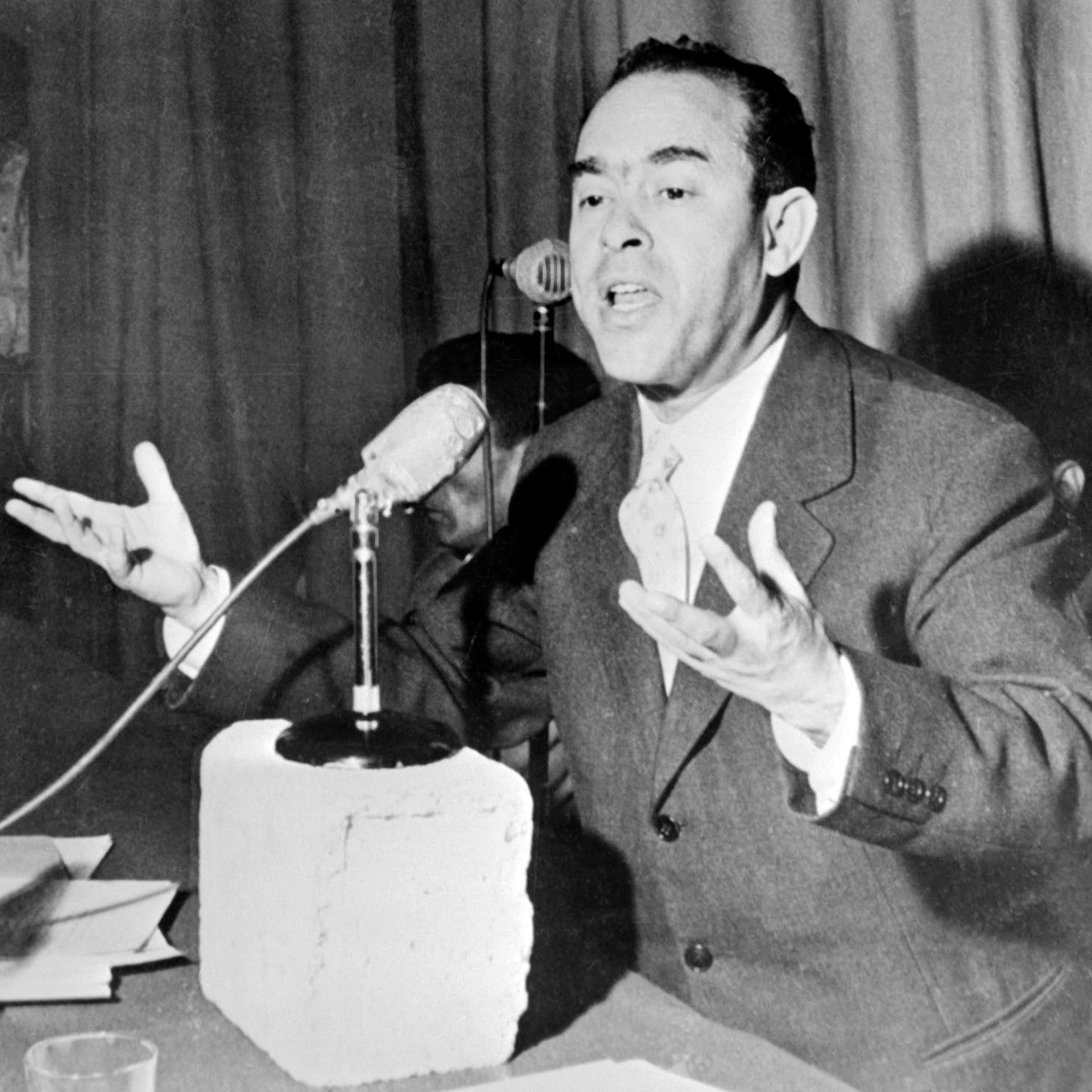 L'image montre un homme en train de s'exprimer avec enthousiasme lors d'une conférence ou d'un discours. Il est vêtu d'un costume et parle devant un microphone, gesticulant avec les mains pour accentuer ses propos. Sur la table devant lui, on peut voir un bloc de béton et quelques documents. En arrière-plan, une autre personne semble prendre des photos ou enregistrer. L'ambiance paraît sérieuse, soulignant l'importance de son discours.