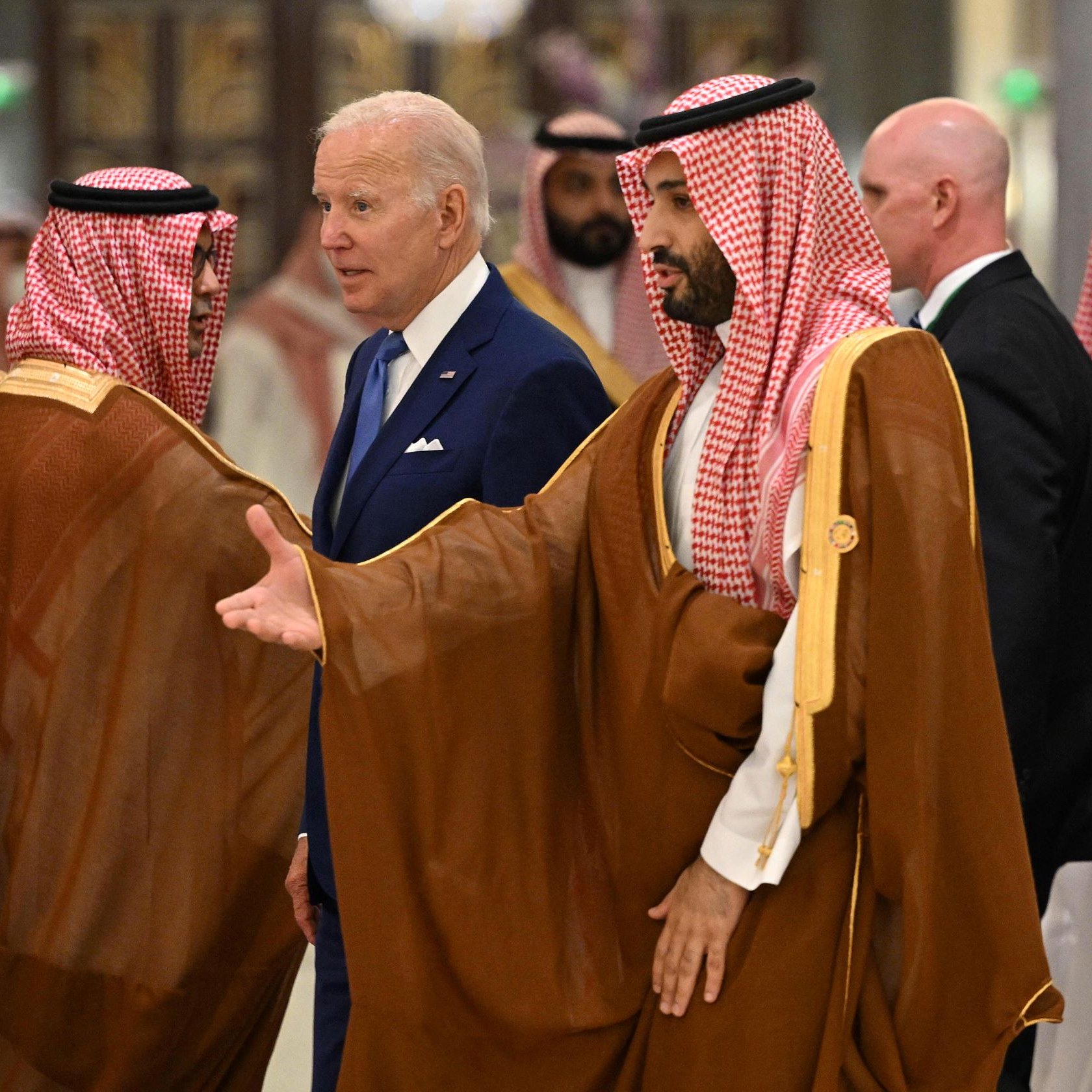 L'image montre un groupe d'hommes dans un environnement intérieur élégant. Au centre, un homme en costume bleu semble converser avec d'autres hommes vêtus de vêtements traditionnels arabes, notamment des thobes et des headdresses en keffieh. Ils sont entourés d'un décor luxueux, probablement dans un palace ou un lieu officiel. Les expressions des hommes montrent une interaction diplomatique.