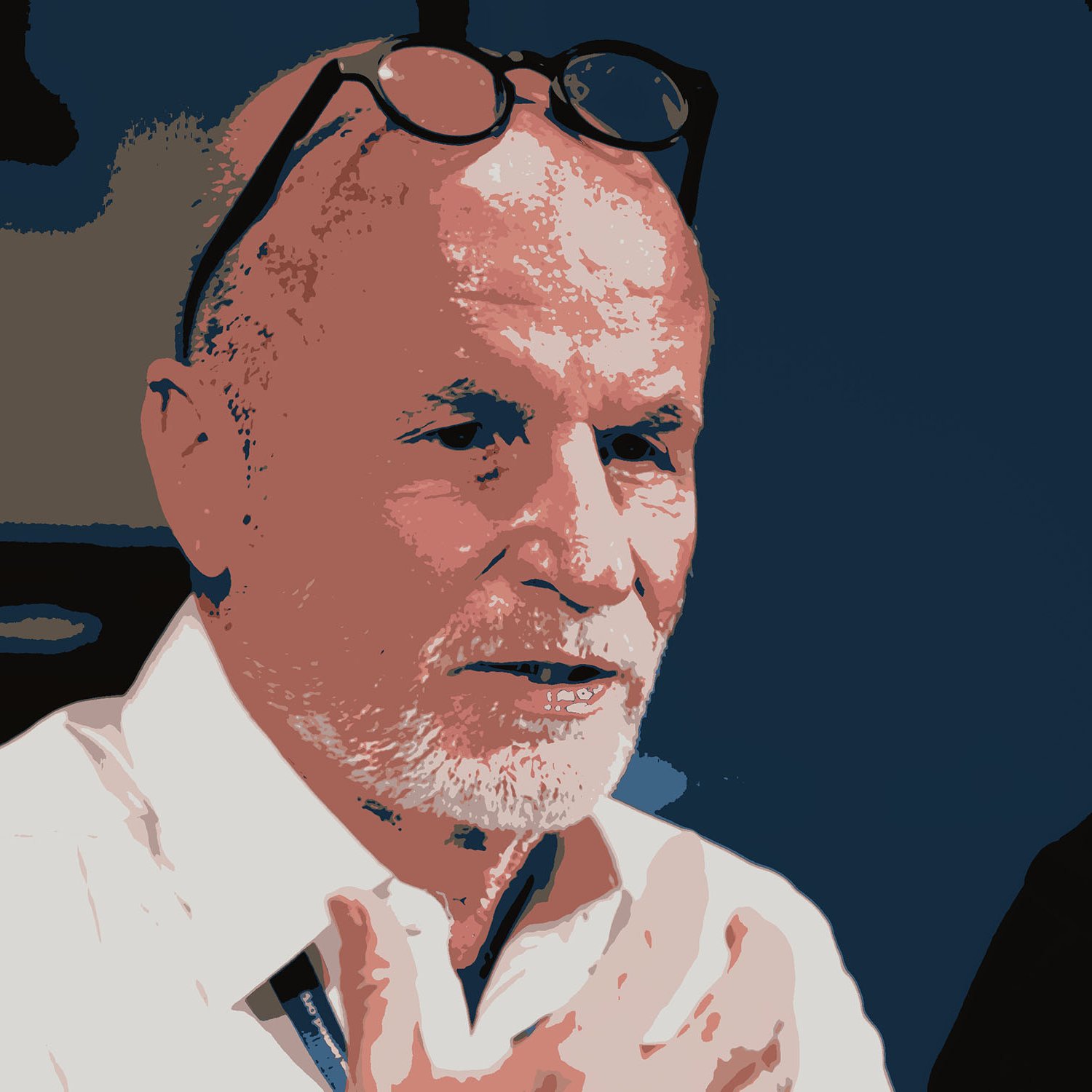 L'image représente un homme avec des cheveux grisonnants et une barbe. Il porte une chemise blanche et des lunettes posées sur sa tête. Son expression semble sérieuse et il a l'air de parler ou d'écouter attentivement. Le fond est de couleur sombre, ce qui met en valeur son visage.