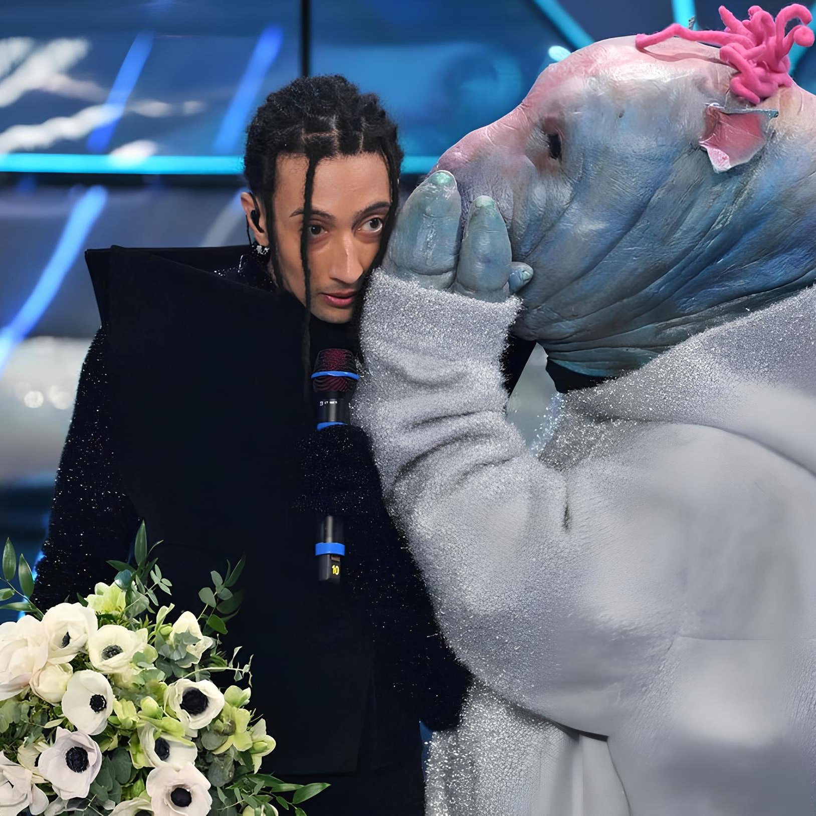 در این تصویر، یک فرد با ظاهری خاص و لباس جذاب در حال گفتگو با یک موجود خیالی است که صورتش شبیه به یک حیوان با پوست آبی و جزئیات رنگی است. این موجود همچنین لباسی پشمی و براق به تن دارد. فرد در حال صحبت ظاهراً شوکه یا متفکر به نظر می‌رسد و در دستش دسته‌گلی زیبا دارد. در پس‌زمینه، نوارهای نوری و طراحی مدرن دیده می‌شود که حال و هوای پویا و جالبی به تصویر می‌دهد.