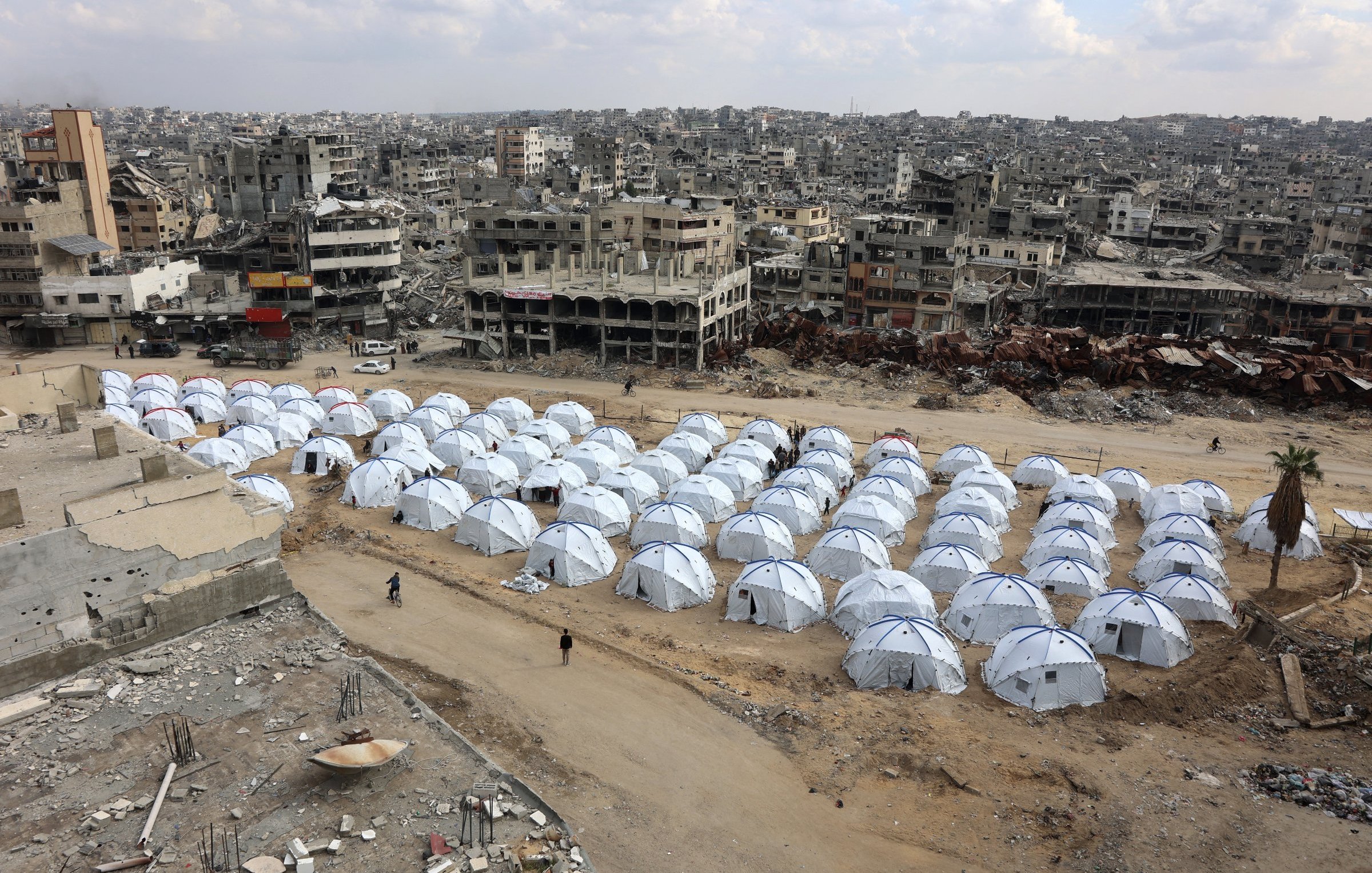 L'immagine mostra un'ampia area con numerosi tende bianche disposte in file ordinate, che rappresentano un campo per sfollati. Sullo sfondo si possono vedere edifici distrutti e in rovina, segno di conflitti o disastri. Il cielo è nuvoloso, contribuendo a un'atmosfera desolante. La scena evidenzia il contrasto tra le tende temporanee, che offrono riparo, e l'ambiente urbano devastato circostante.