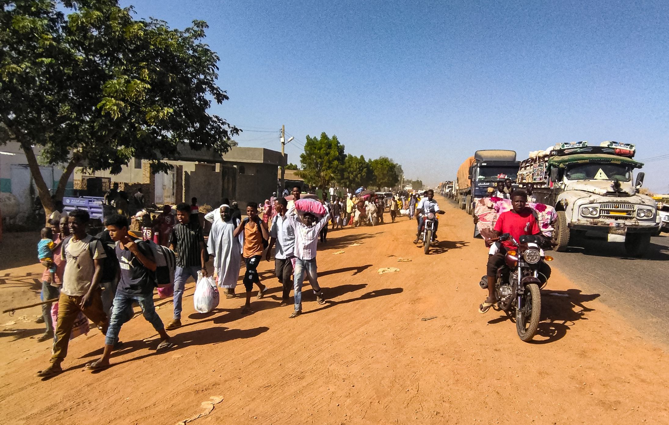 L'immagine mostra una scena di vita quotidiana lungo una strada in un ambiente urbano o semi-urbano. Si notano molte persone che camminano lungo il lato della strada, alcune con sacchi o beni in mano, e altre che sembrano discutere tra di loro. Sul lato opposto della strada ci sono veicoli in transito, tra cui camion e motociclette. La vegetazione circostante è presente, con alcuni alberi che offrono ombra. Il terreno appare polveroso, e l'atmosfera complessiva sembra vivace e movimentata.