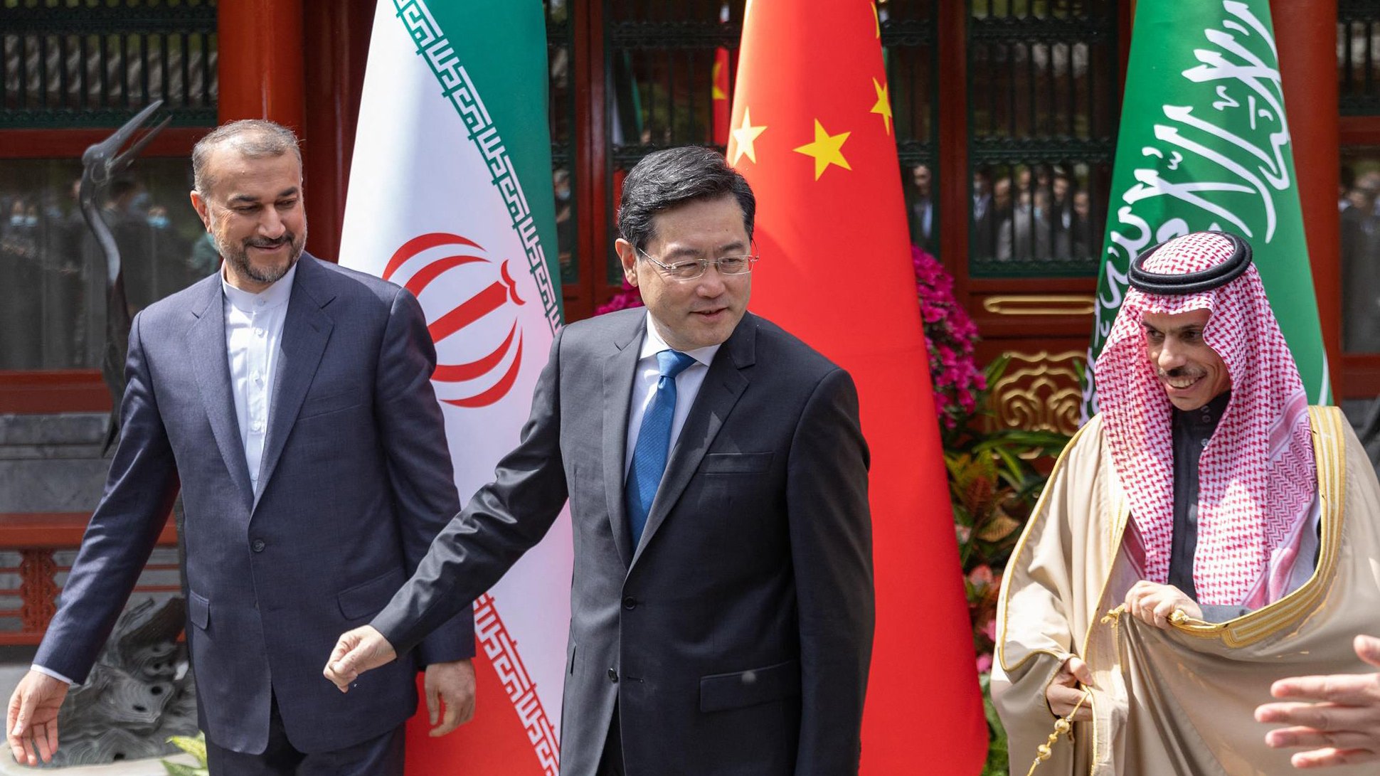 La imagen muestra a tres hombres en un ambiente formal, posiblemente en una reunión diplomática. A la izquierda, un hombre vestido con un traje oscuro parece estar sonriendo ligeramente. En el centro, otro hombre con una corbata azul parece estar en movimiento, mientras que a la derecha, un tercer hombre vestido con una túnica clara y una capa está sonriendo abiertamente. Detrás de ellos se pueden ver banderas de Irán, China y Arabia Saudita, lo que sugiere que esta reunión involucra a estos tres países. El entorno parece ser elegante y bien cuidado.