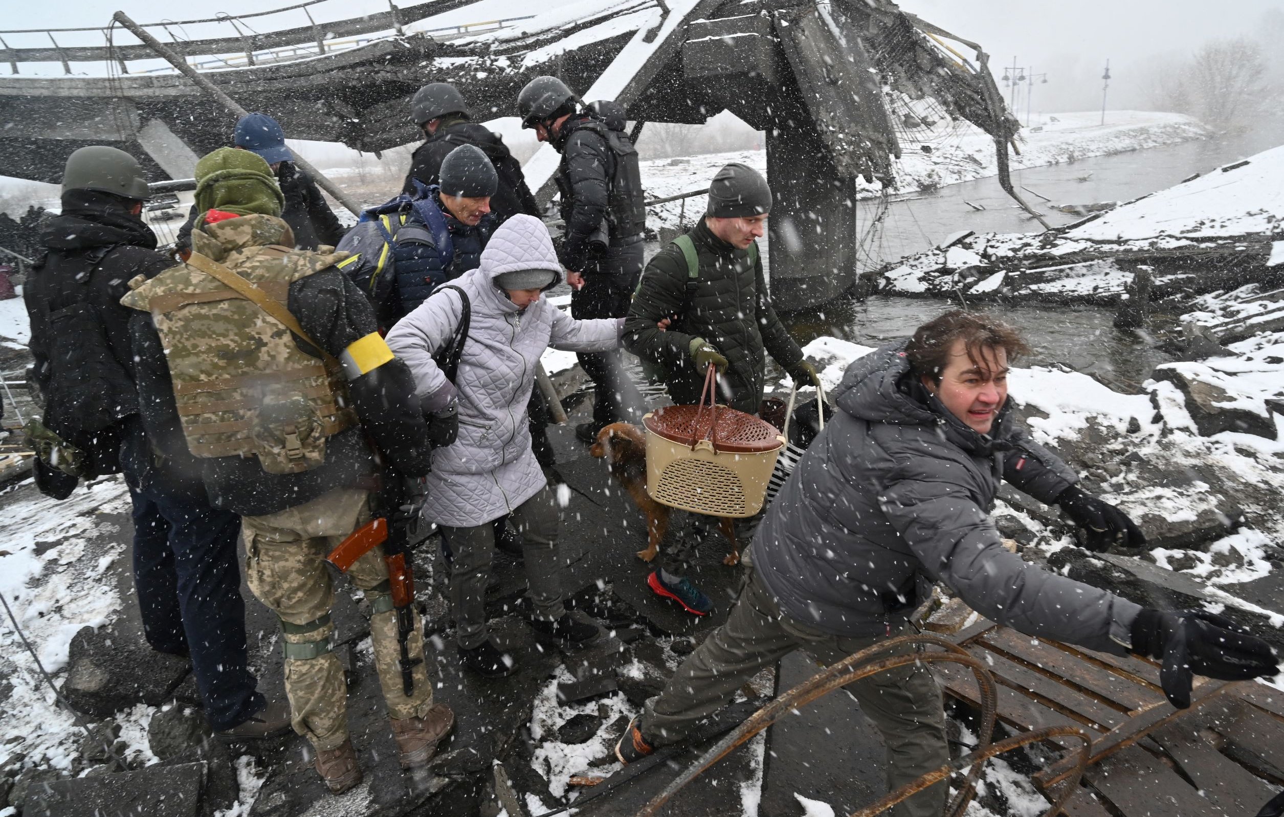 La imagen muestra un grupo de personas en un entorno destruido, posiblemente tras un conflicto o desastre. Hay varios individuos vestidos con ropa de abrigo, algunos de ellos parecen ser militares. La escena se desarrolla en un lugar cubierto de nieve, con escombros visibles y una estructura dañada en el fondo. Algunas personas están ayudando a otros, sugiriendo un intento de rescate o evacuación. El ambiente es tenso y hay un sentimiento de urgencia en la acción de los que están presentes. La nieve está cayendo, lo que agrega a la dureza de la situación.