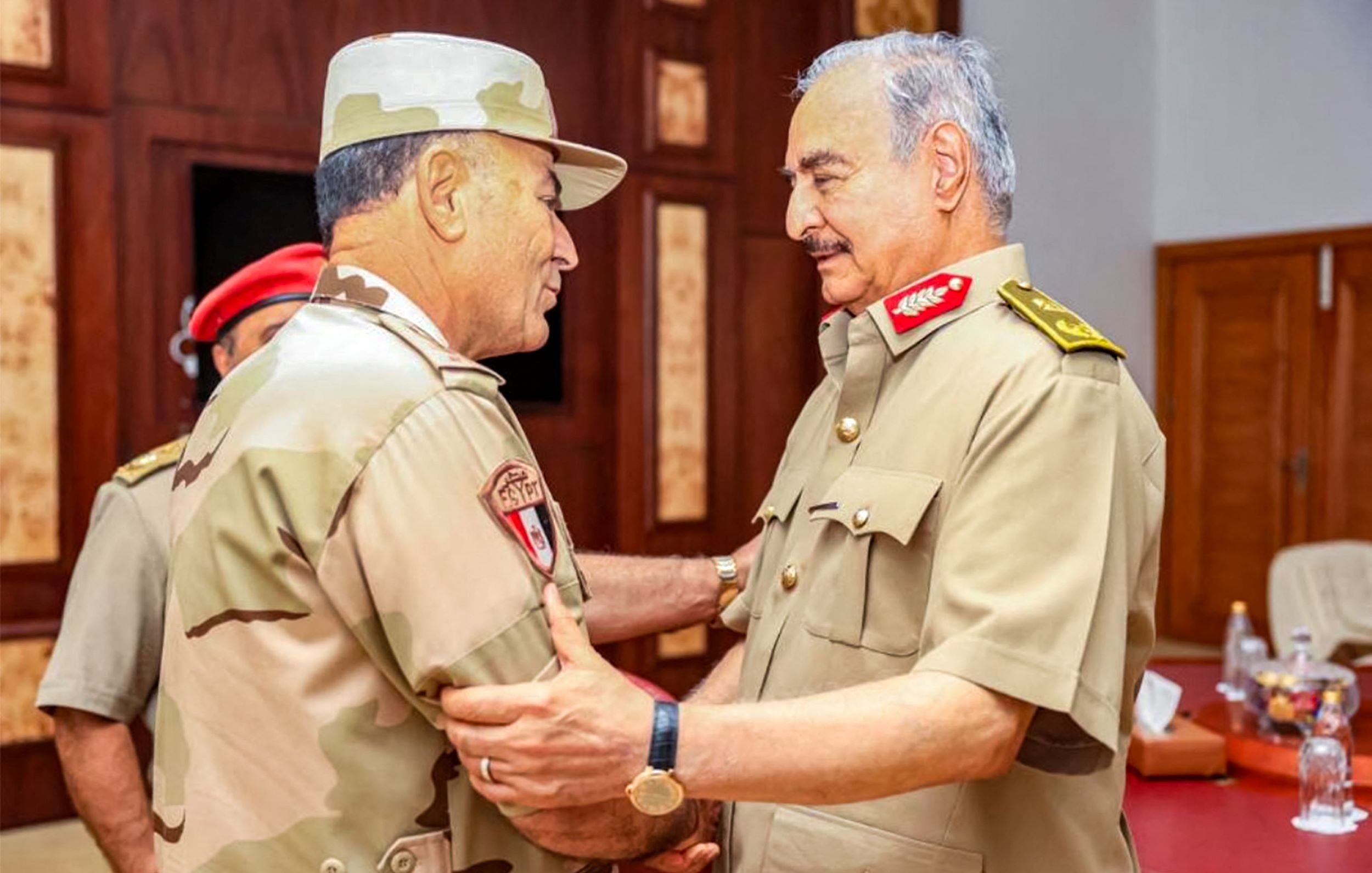 L'image montre deux militaires en uniforme, se tenant face à face et en train de discuter. L'un d'eux porte un chapeau militaire et un uniforme beige avec des motifs camouflage, tandis que l'autre est habillé en uniforme militaire beige et porte des décorations sur son épaule. Ils semblent avoir une conversation amicale et posent dans un environnement intérieur, avec des tables et des chaises visibles en arrière-plan. L'atmosphère semble informelle, suggérant une réunion ou un échange entre officiers.
