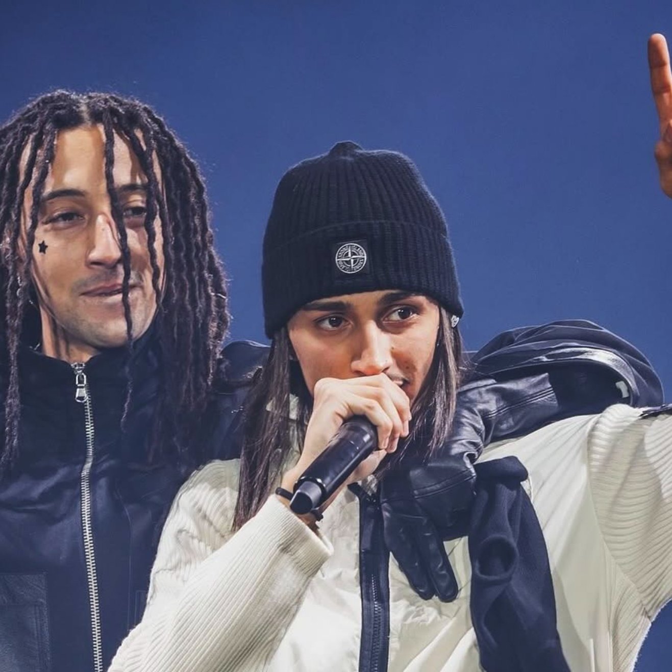 Due artisti sul palco: uno con capelli ricci e l'altro con un cappello nero, entrambi in abbigliamento sportivo.