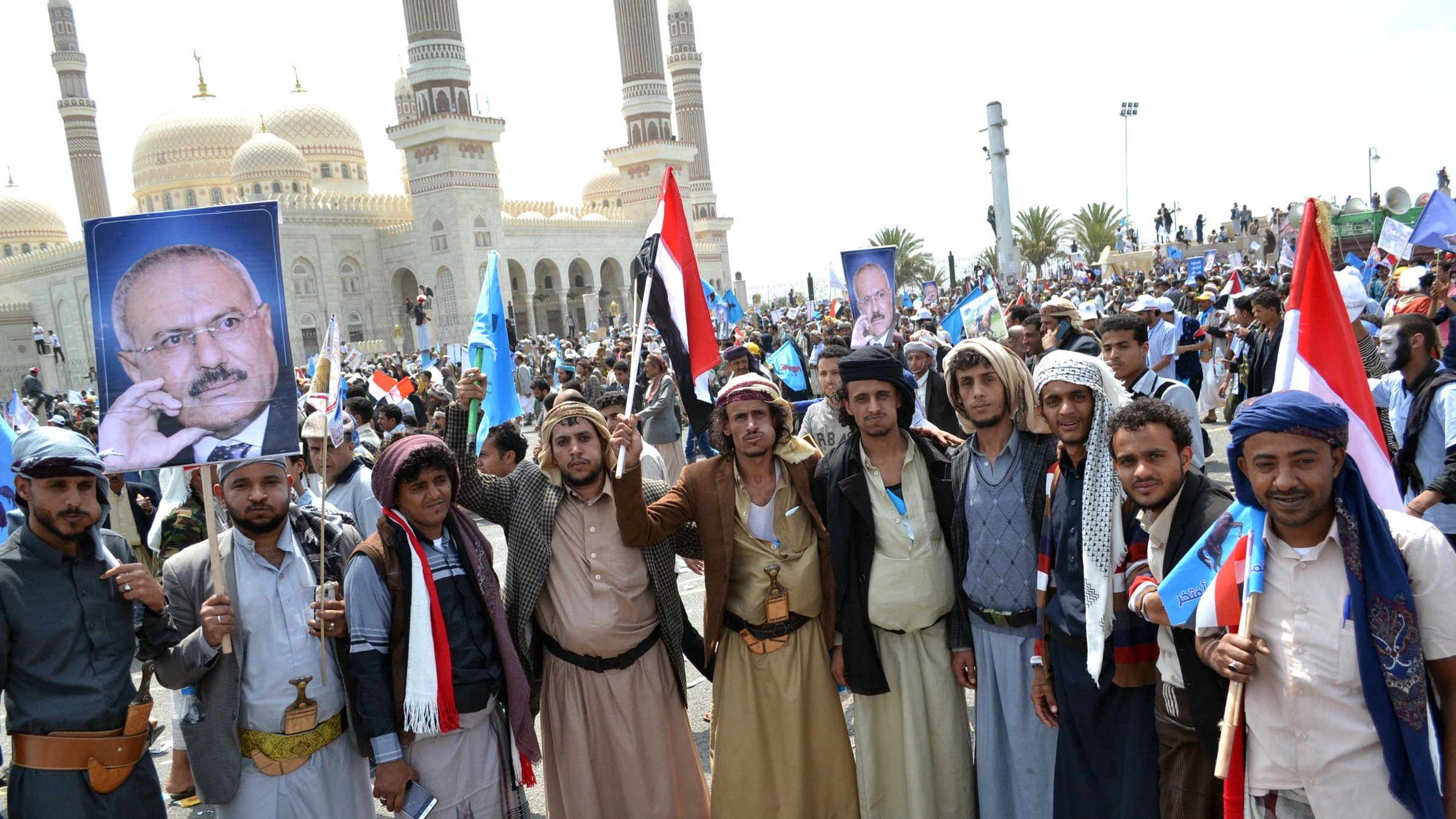 L'image montre un groupe de personnes rassemblées lors d'une manifestation. Elles portent des vêtements traditionnels et tiennent des drapeaux ainsi que des pancartes, y compris une qui affiche un portrait. En arrière-plan, on peut voir une grande mosquée ou un bâtiment avec des dômes, suggérant que l'événement a lieu dans un environnement urbain. L'atmosphère semble festive et engagée, avec une forte mobilisation des participants.