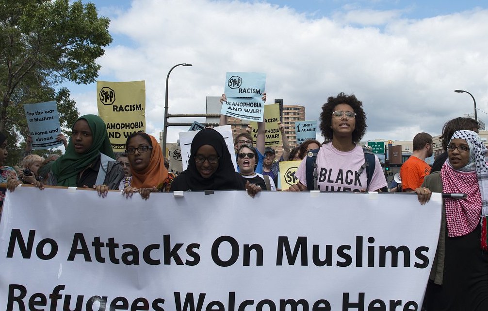 في الصورة، تظهر مجموعة من الأشخاص المشاركين في مظاهرة. يحمل المتظاهرون لافتات مكتوب عليها عبارات تدعو للتضامن مع المسلمين وترحب باللاجئين. يرتدي بعض النساء الحجاب، وتظهر تعابير الحماس والتأييد على وجوههم. السماء تبدو غائمة قليلاً، مما يضفي توتراً على الأجواء، وفي الخلفية يمكن رؤية مب buildings وشعارات تعبر عن رفض العنصرية والتمييز.