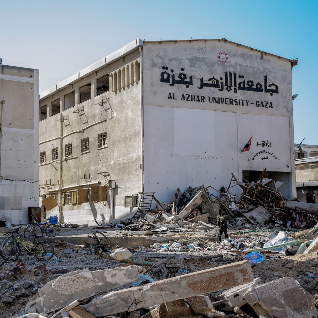 L'image montre un paysage de destruction, probablement à Gaza. On aperçoit un bâtiment, vraisemblablement l'Université Al-Azhar, dont la façade est endommagée. Des débris et des gravats jonchent le sol, indiquant des destructions significatives. Deux personnes à vélo se déplacent parmi les débris, ce qui souligne la difficulté de la situation. Le mur du bâtiment est partiellement effondré, et des inscriptions en arabe sont visibles. L'atmosphère de l'image est sombre, révélant les conséquences d'un conflit.