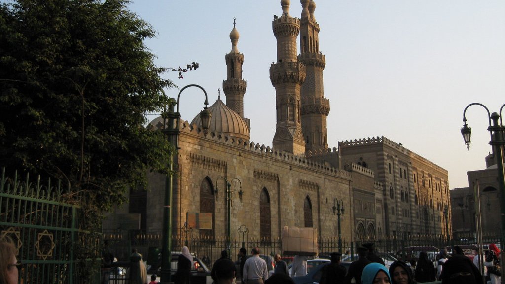 L'image montre un vaste bâtiment architectural au style islamique, probablement une mosquée, avec des minarets majestueux s'élevant vers le ciel. On peut voir des personnes en tenue traditionnelle, certaines portant le hijab. En arrière-plan, la lumière du soleil est douce, créant une atmosphère paisible. Les détails architecturaux de la façade en pierre ajoutent une touche historique à la scène. Des arbres et des lampadaires embellissent également l'environnement.