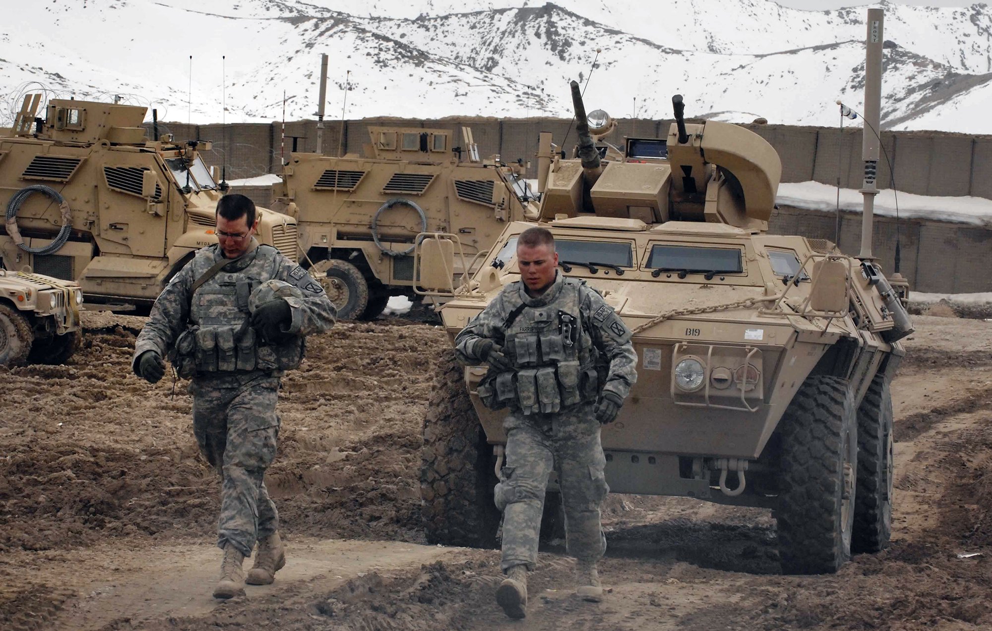 L'image montre deux soldats marchant dans un environnement rocailleux, probablement dans une zone militaire. Ils portent des uniformes camouflage et des gilets tactiques. En arrière-plan, on peut voir des véhicules blindés semblant robustes, probablement utilisés pour des opérations militaires, avec des montagnes enneigées en toile de fond. L'atmosphère semble sérieuse et empreinte d'une certaine rigueur militaire.
