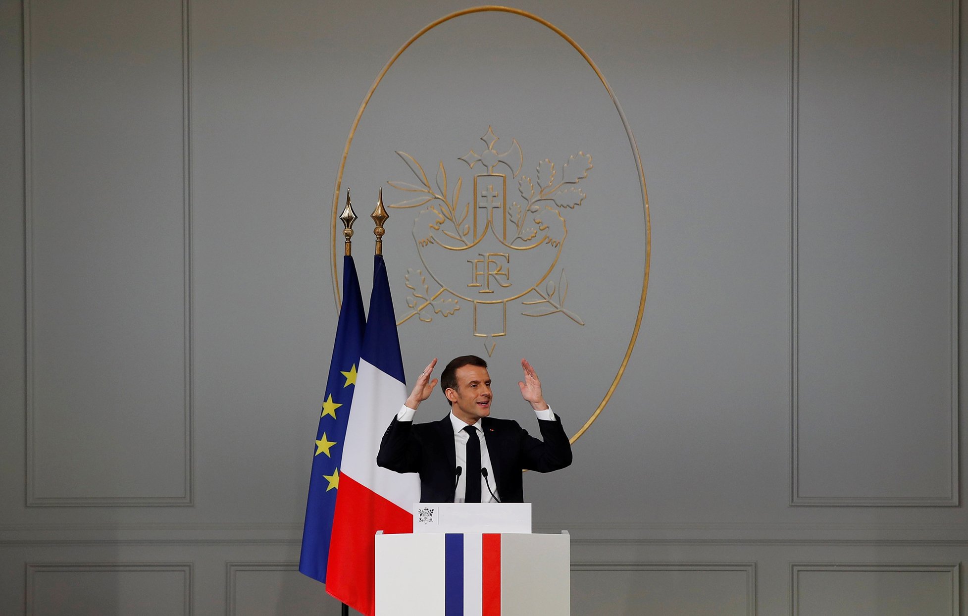 در این تصویر، یک مرد در حال سخنرانی است. او در جلوی یک تریبون ایستاده و دست‌هایش را به سمت بالا برده است. در پشت او، نماد رسمی فرانسه وجود دارد که شامل دو پرچم می‌باشد؛ یکی پرچم فرانسه و دیگری پرچم اروپایی. دیوار پشت او ساده و به رنگ خاکستری است و نماد جمهوری فرانسه در مرکز قرار دارد. این صحنه نشان‌دهنده یک رویداد رسمی یا کنفرانس مطبوعاتی است.