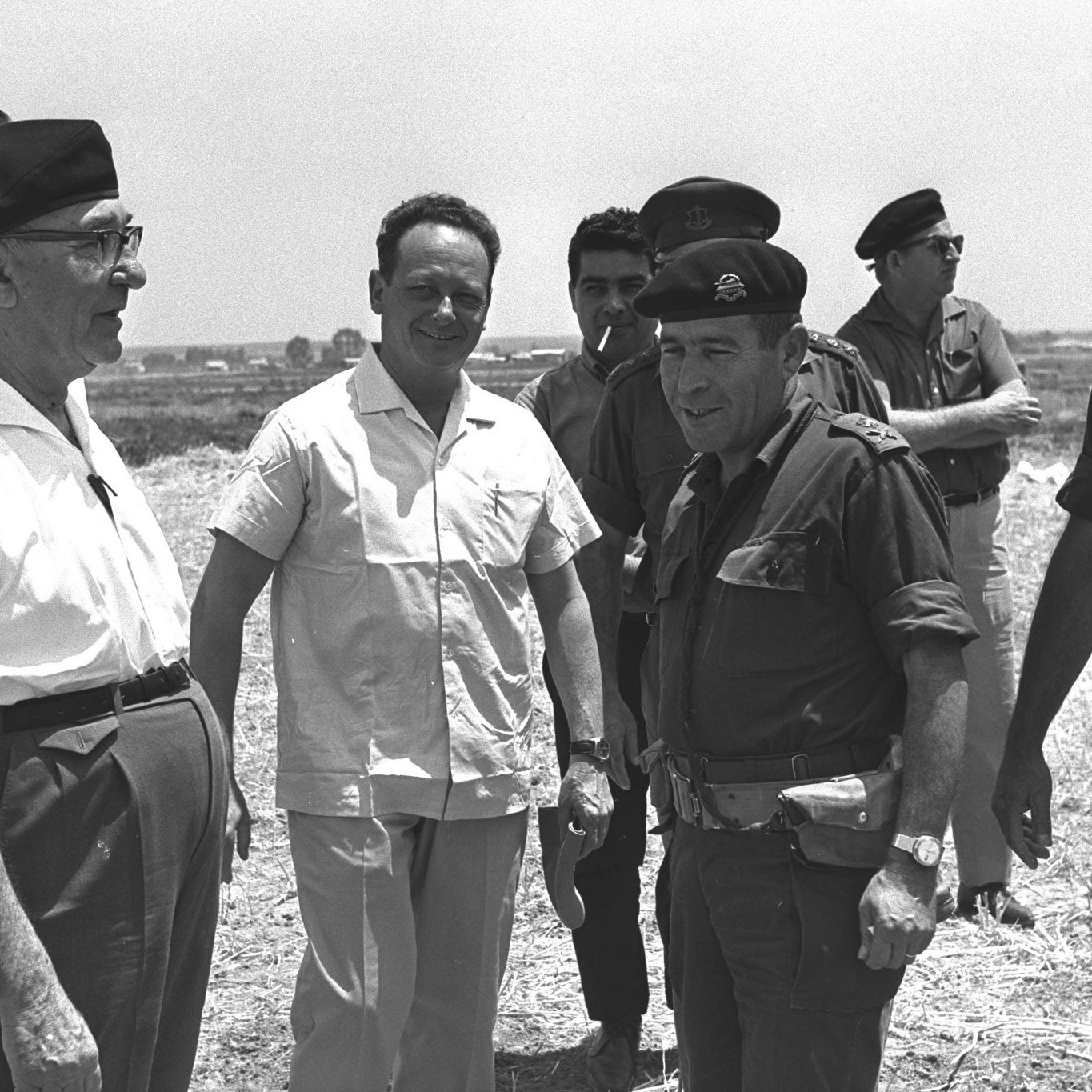 L'image montre un groupe d'hommes en train de discuter à l'extérieur. Certains portent des uniformes militaires, tandis que d'autres sont habillés de façon plus décontractée, comme des chemises blanches. L'atmosphère semble informelle, et les participants échangent vraisemblablement des opinions ou des informations dans un cadre ensoleillé. En arrière-plan, on peut apercevoir un paysage aride.