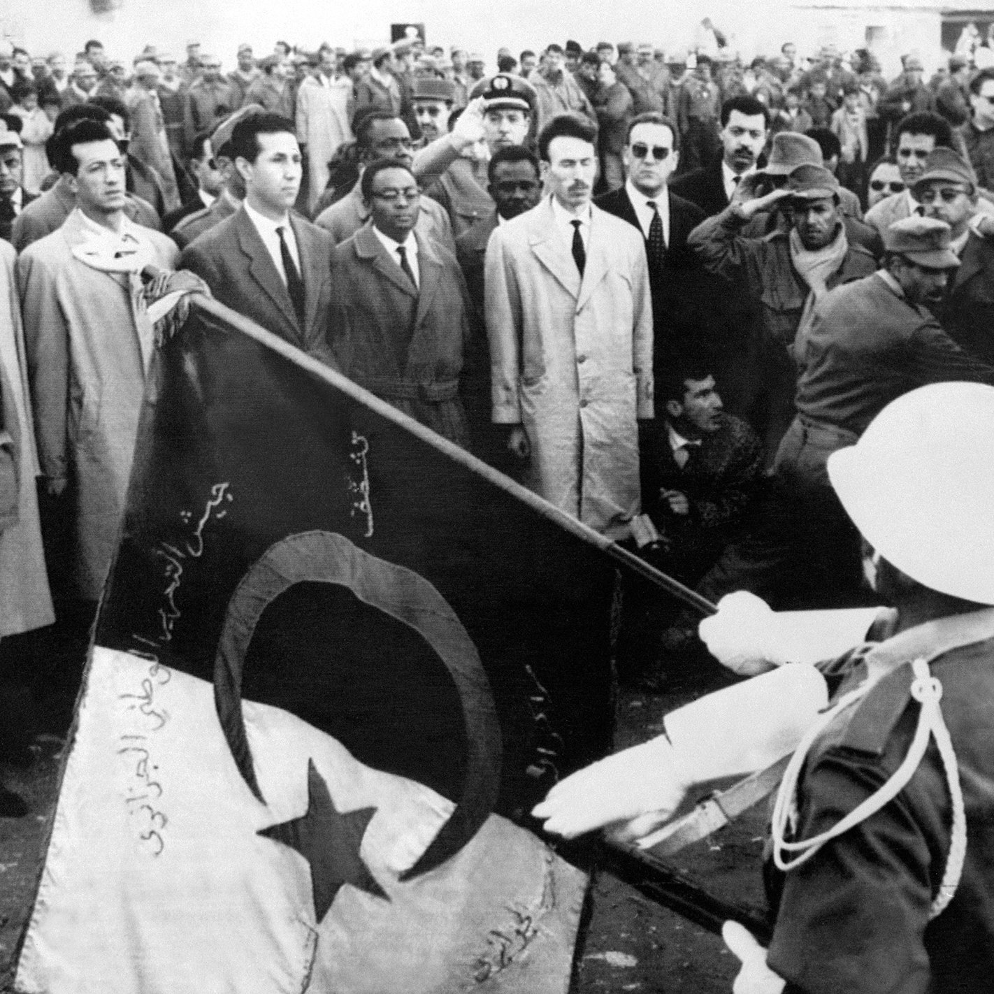 Cette image en noir et blanc semble représenter un événement historique, probablement lié à la décolonisation ou à un moment significatif dans l'histoire de l'Algérie. On peut voir un groupe de personnes rassemblées, avec des hommes en tenue civile et militaire. Au premier plan, des personnages saluent tandis que des soldats présentent un drapeau. L'atmosphère semble chargée d'émotion et de solennité, indiquant un moment important qui pourrait symboliser la fierté nationale ou la lutte pour l'indépendance.
