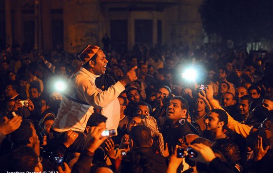 L'image montre une grande foule rassemblée dans une ambiance nocturne. Un homme au centre, visiblement passionné, est en train de parler ou de s'exprimer avec intensité. Les visages de la foule sont éclairés par des lumières, peut-être des téléphones portables ou des caméras. Les expressions des gens varient entre l'engagement, la curiosité et l'émotion, suggérant un moment de rassemblement ou de protestation. L'ensemble dégage une atmosphère vibrante et dynamique.