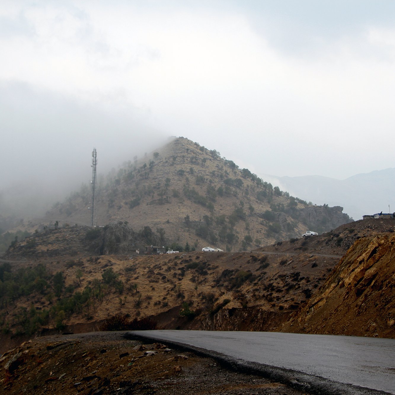 L'image montre un paysage montagneux avec des collines voilées par le brouillard. En arrière-plan, une structure de télécommunication est visible au sommet d'une colline. La route qui serpente est goudronnée, et le terrain environnant présente des nuances de terre brune et de verdure. L'atmosphère semble sombre et mystérieuse, suggérant une journée nuageuse et humide.