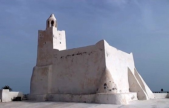 L'image montre un bâtiment de couleur blanche, typiquement de style architectural méditerranéen. Il semble être une petite structure, possédant une forme irrégulière avec des murs épais et angulaires. À son sommet, il y a une cloche ou un petit clocher. L'arrière-plan est plutôt clair, avec un ciel bleu ou nuageux qui met en valeur la luminosité de l'édifice. Ce type de construction pourrait évoquer des lieux historiques ou religieux, typiques de certaines régions côtières.