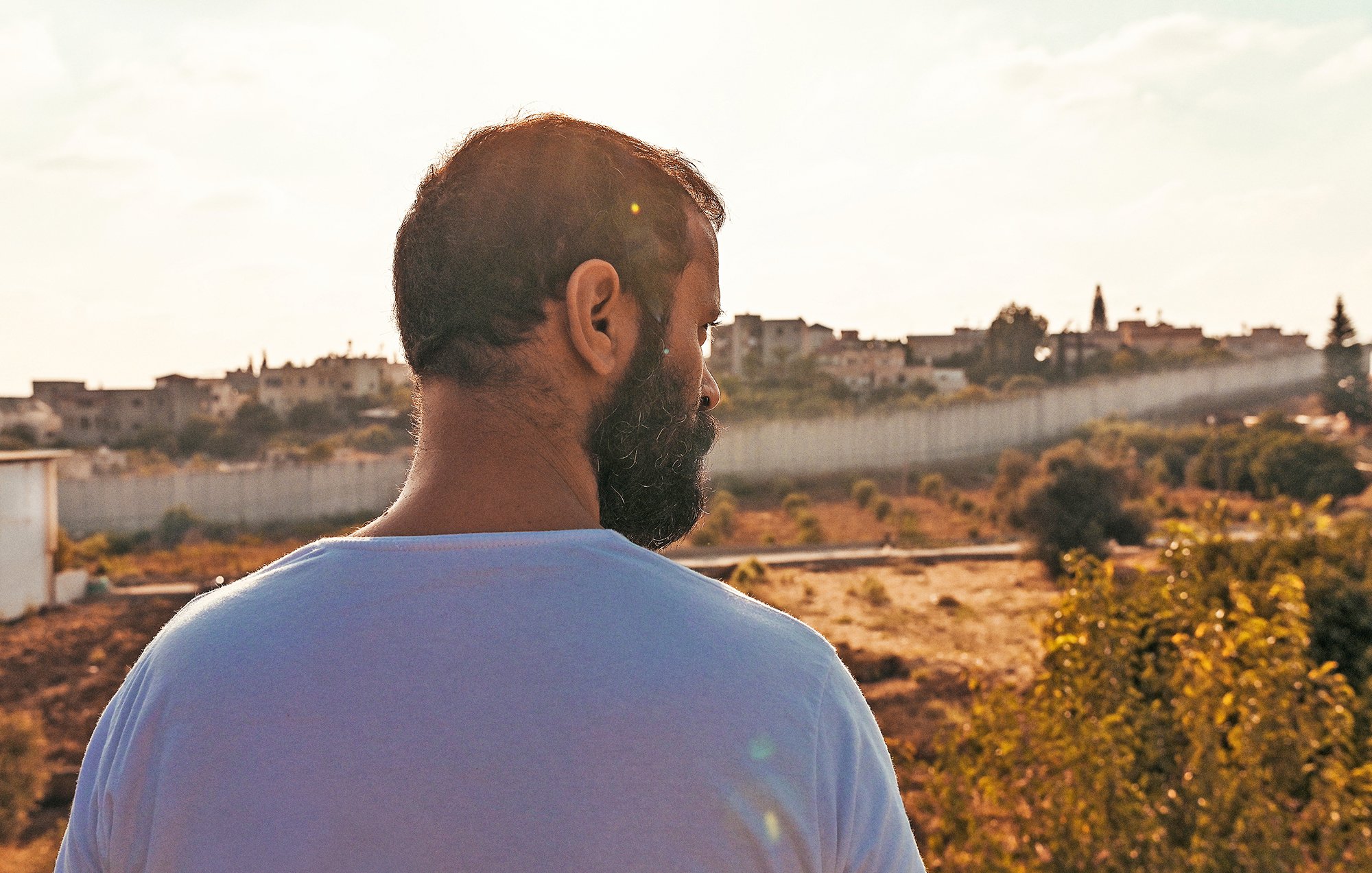 L'image montre un homme vu de dos, contemplant un paysage. Il a une barbe et porte un t-shirt blanc. En arrière-plan, on aperçoit des bâtiments sur une colline, flanqués d'une barrière ou d'un mur. La lumière est douce, créant une atmosphère paisible, et le ciel présente quelques nuages. Le terrain est principalement sec avec des touches de verdure autour.