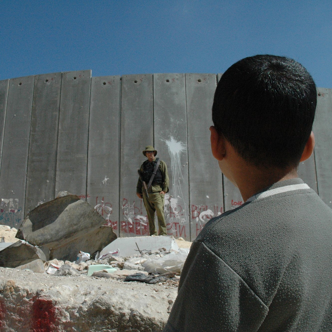 L'image montre un jeune garçon observant un soldat qui se tient devant un mur en béton. Le mur est marqué par des graffitis, suggérant un environnement de conflit ou de tension. Des débris sont dispersés au sol, et le ciel est dégagé, ce qui contraste avec la gravité de la scène. Le regard du garçon semble interrogateur ou curieux face à la situation qui l'entoure.