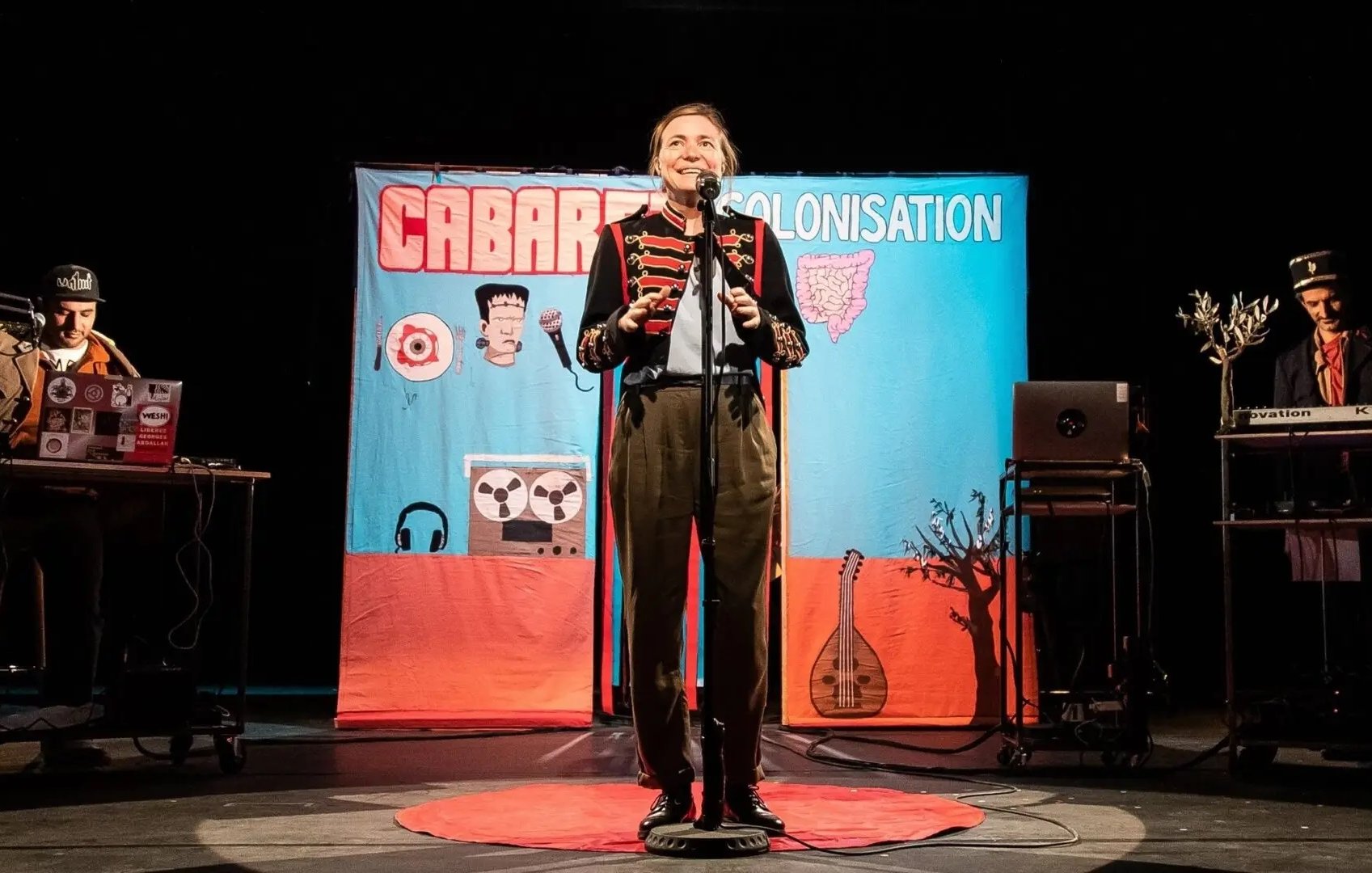 L'image montre une scène de spectacle, probablement un cabaret. Au centre, un artiste souriant est sur scène avec un micro, habillé de manière théâtrale. En arrière-plan, un grand panneau coloré affiche le mot "CABARET" et "COLONISATION", suggérant un thème spécifique. De chaque côté, on aperçoit des musiciens ou des techniciens avec des instruments ou des équipements, contribuant à l'atmosphère festive et engageante. Le sol est orné d'un tapis rouge, ajoutant une touche de mise en scène au spectacle.