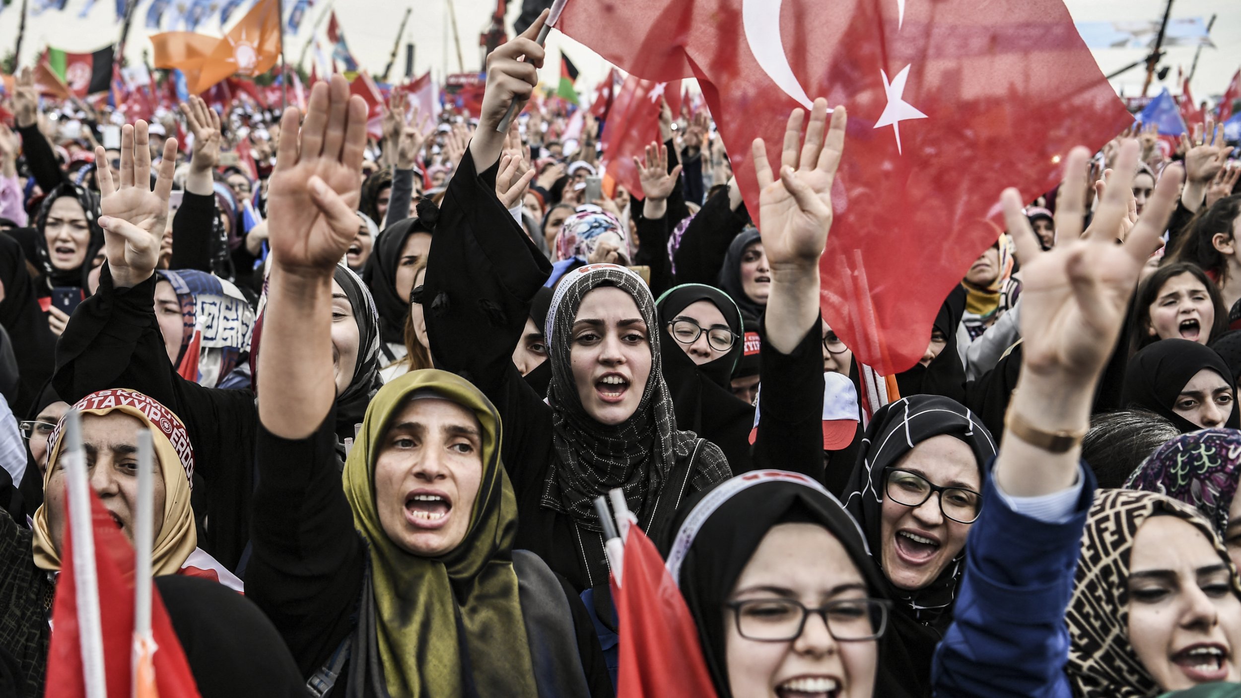 L'image montre une grande foule de personnes, principalement des femmes, qui sont rassemblées lors d'un événement ou d'une manifestation. Elles expriment leur enthousiasme et leur soutien en levant des drapeaux, dont certains arborent le symbole de la Turquie. Les participantes portent des vêtements traditionnels ou des foulards, et certaines semblent chanter ou crier des slogans. L'ambiance est dynamique et remplie d'énergie collective, suggérant une atmosphère de célébration ou de revendication.